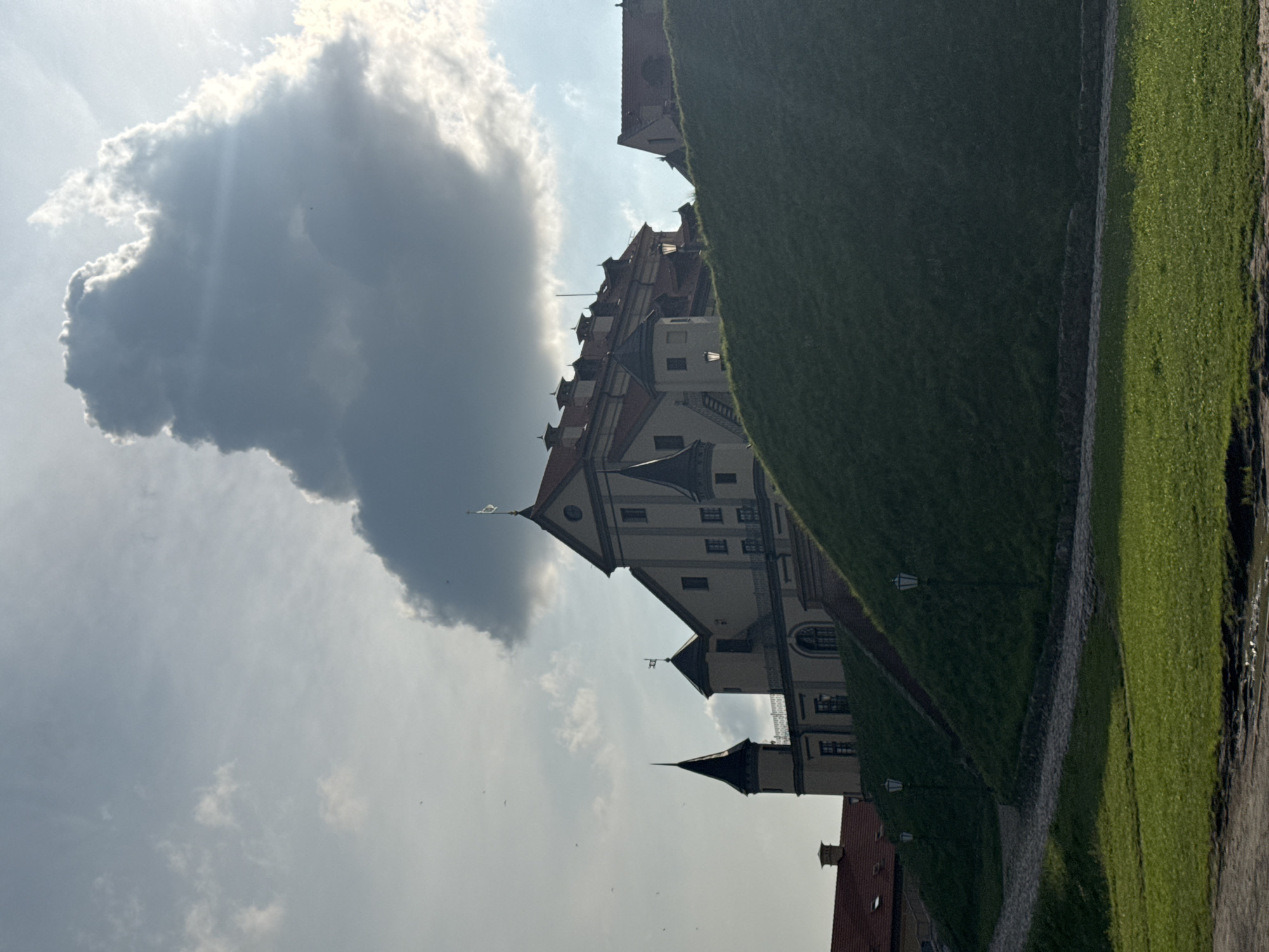 This image depicts a view of the Nesvizh Castle in Nesvizh, Belarus. The castle, a large, pale yellow building with red tiled roofs and several conical towers, is situated atop a grassy mound. A large, dark cloud dominates the sky above the castle. The foreground shows a winding cobblestone path and a well-maintained grassy area. Several lamp posts are visible along the path and on the mound. The lighting suggests it is daytime, with sunlight filtering through gaps in the clouds. The overall scene is serene and historical, highlighting the architecture and surrounding landscape of the castle.