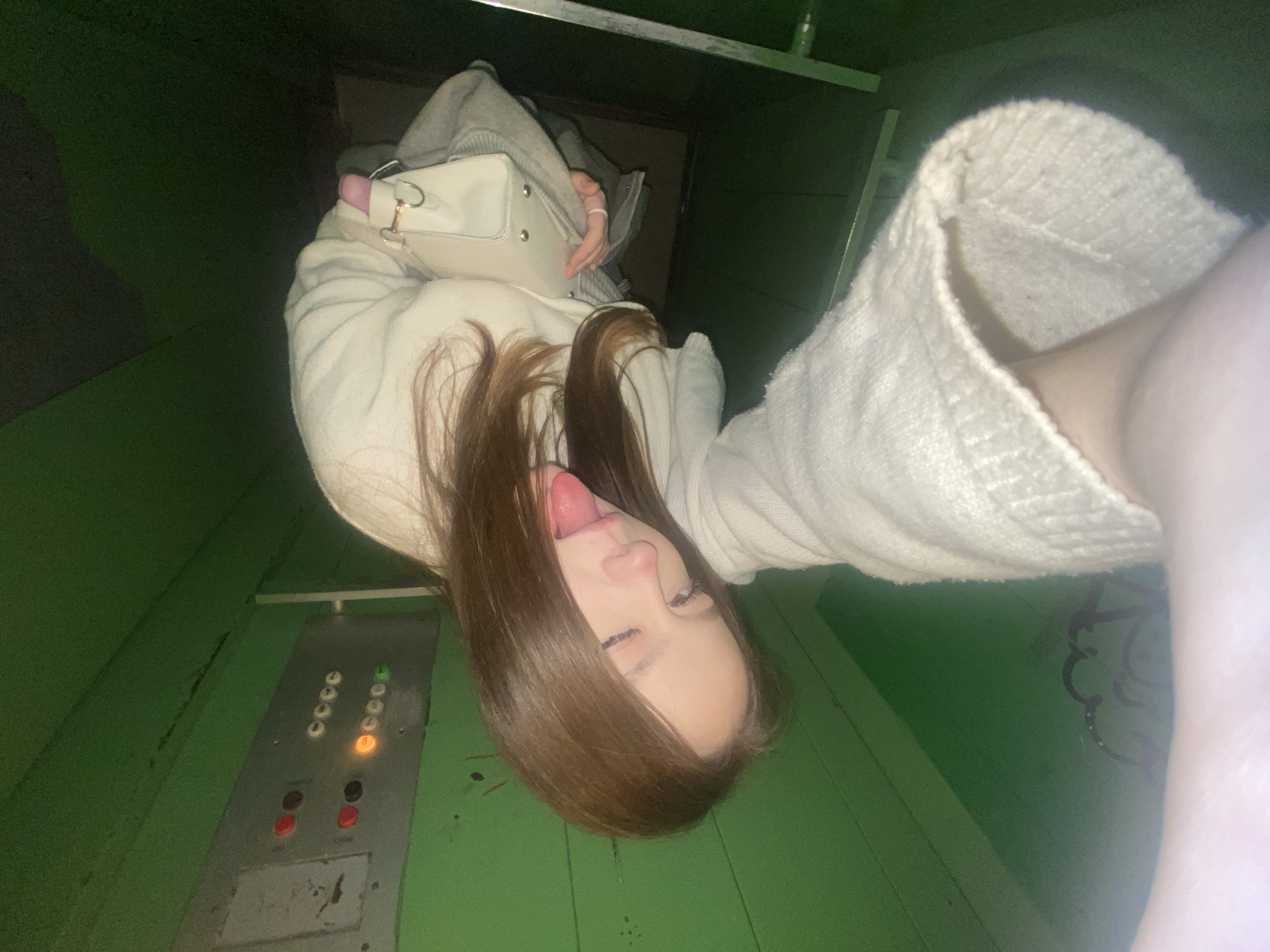 The image captures a playful selfie of a young person inside what appears to be an elevator in Chaikovsky, Russia. The individual is oriented upside down in the frame, featuring long brown hair cascading downwards. They are wearing a light-colored (possibly white or cream) knitted sweater or sweatshirt and holding a white handbag with gold-toned hardware. The person is sticking out their tongue and winking one eye, conveying a casual and cheeky expression.
The setting is an enclosed space with distinctive green walls, suggesting an older elevator. An elevator control panel is visible to the person's left (when the image is viewed as displayed). The panel has two columns of round buttons; some are illuminated, with one button displaying an orange light, likely indicating a floor selection or current position. Below these, there are two larger red circular buttons. The green walls show signs of wear, including scratches and some faint, dark, abstract graffiti on the right side. The lighting within the elevator is artificial and somewhat dim.