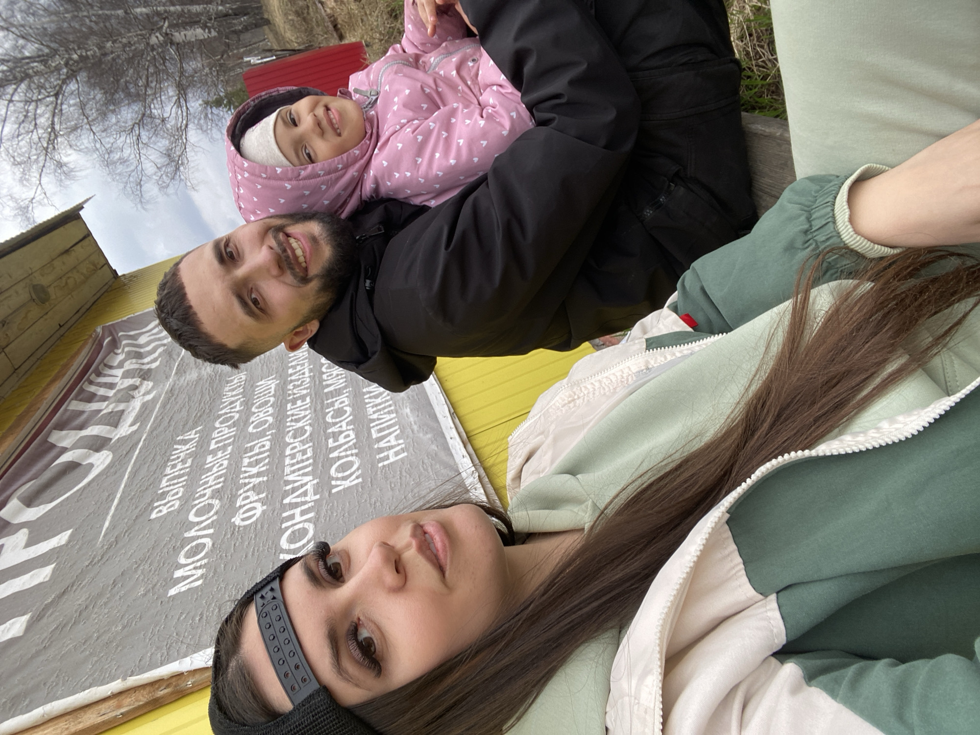 The image shows three people: a woman, a man, and a young girl. The woman is in the foreground, lying down and facing the camera. She has long dark hair and is wearing a dark headband and a green and white jacket. The man is behind her, holding the girl in his arms. The girl is wearing a pink jacket with white hearts. The background includes a sign with text in Russian, a building with a yellow facade and a red roof, and some trees. The location is Zhigalki, Russia.