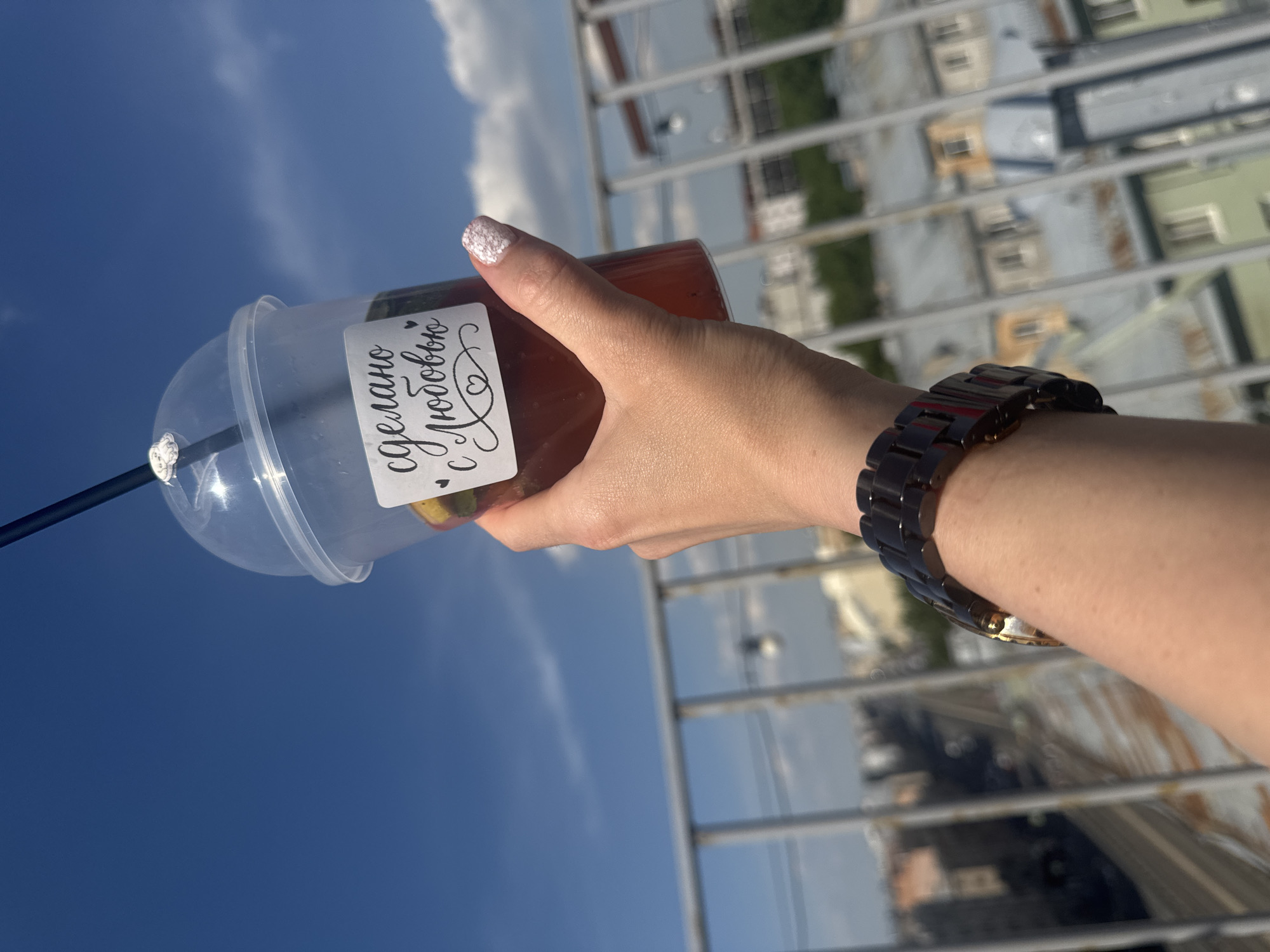 A person's hand is holding a clear plastic cup filled with a reddish-brown beverage and ice, against a bright blue sky with scattered clouds. The cup has a white label with black cursive text that reads "сделано с любовью" (made with love) and a small heart. A black straw protrudes from the top of the cup, secured with a white ring. The person is wearing a dark, multi-segmented wristwatch on their left wrist. In the background, out of focus, are rooftop views of buildings in Saint Petersburg, Russia, with grey tiled roofs and some visible windows and balconies. A metal railing runs horizontally across the mid-ground. The lighting suggests it is daytime, likely sunny.