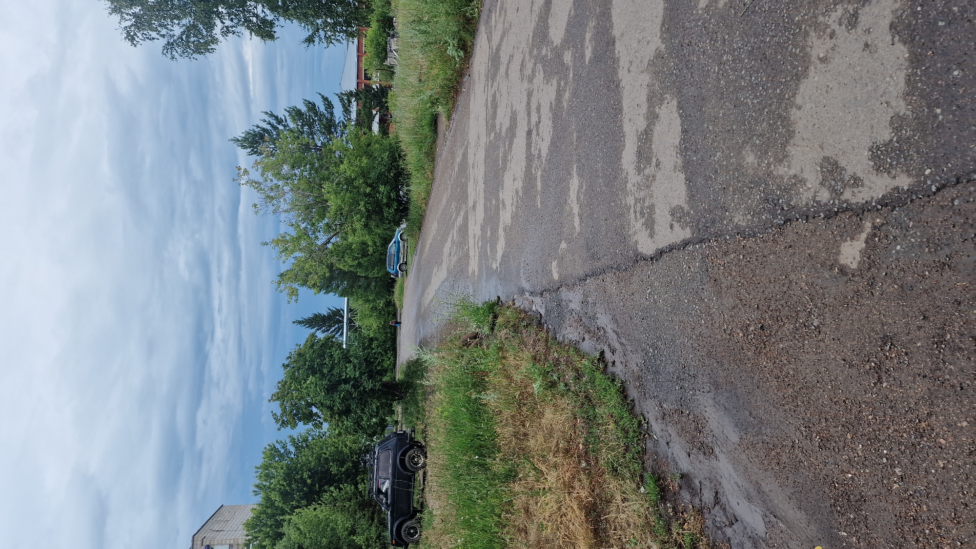 The image depicts an outdoor scene, likely a road or pathway, with a cloudy sky overhead. On the left side of the image, a black SUV is parked on a grassy area next to a gravelly dirt path that appears wet and muddy. Further along the road, a light blue car is visible, and a person is walking in the distance. The road itself is paved with asphalt and shows signs of dampness, with lighter patches indicating where water may have pooled. Lush green trees and foliage line the sides of the road, and in the background, several buildings can be observed, including a brick structure. The overall environment suggests a somewhat rural or suburban setting within a city, possibly during or after a rain shower.
