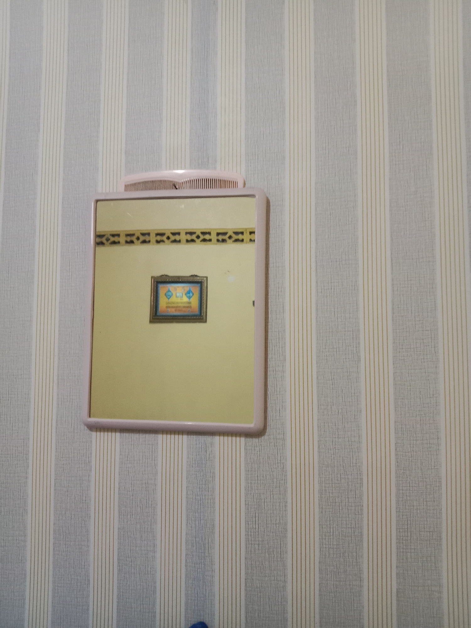A framed picture is reflected in a mirror mounted on a wall with vertical striped wallpaper. The picture frame is dark gold and appears to contain a colorful image with blue and orange elements and text. A pink comb is resting on top of the mirror. The wallpaper features alternating wide light gray stripes and thin yellow stripes. The overall scene suggests an interior setting, possibly a bedroom or bathroom, in Kutosari, Indonesia.