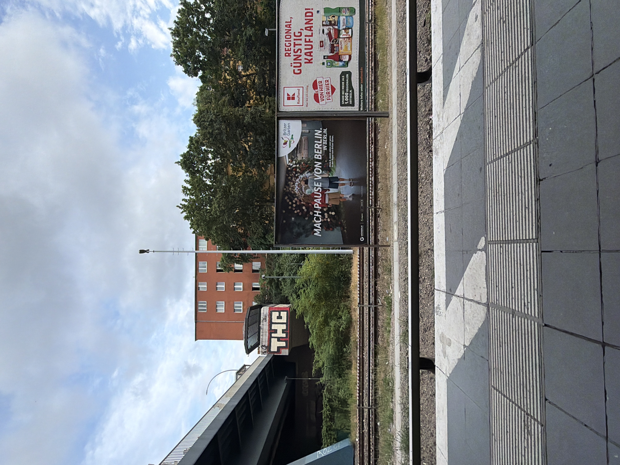 This is a vertical, medium-angle shot of a train station platform in Berlin, Germany. The platform features tactile paving for the visually impaired and a white diagonal line painted on the ground. To the left of the platform are railway tracks and a grassy embankment. Beyond the tracks, a red brick building with multiple windows stands tall, partially obscured by lush green trees. A train with the graffiti "THC" on its front is visible underneath a bridge. Two large billboards are mounted on poles between the building and the train tracks. The top billboard advertises "Kaufland" with the slogan "REGIONAL, GÜNSTIG, KAUFLAND!" and mentions saving on over 1,000 regional articles. The bottom billboard features an image of people enjoying food and the text "MACH PAUSE VON BERLIN. IN BERLIN." It also mentions "Britzer Garten". The sky above is partly cloudy, suggesting daytime. The overall scene depicts a typical urban transit environment with elements of advertising and local attractions.