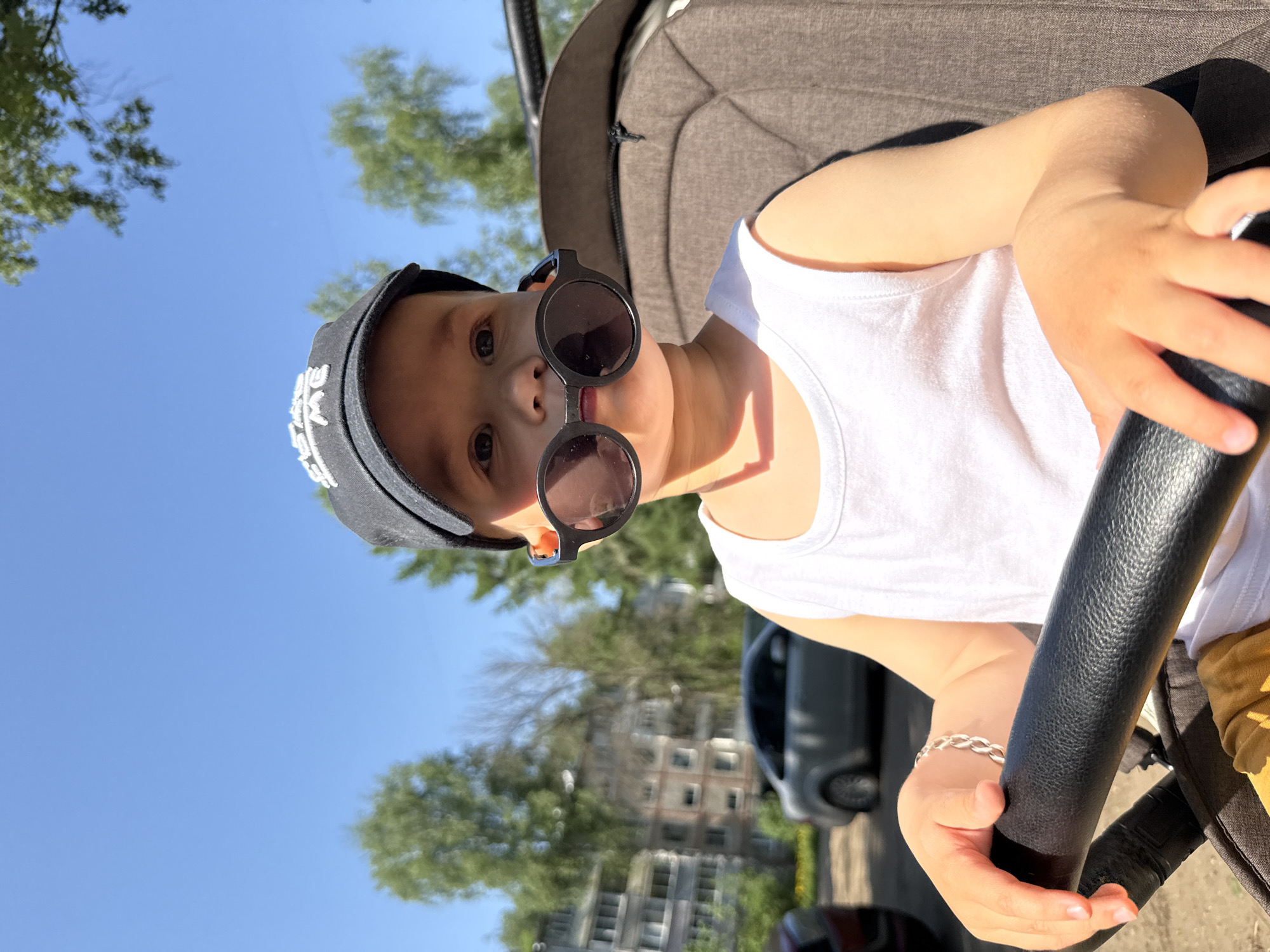 A young child wearing a black baseball cap and round, black sunglasses is in a stroller. The child wears a white tank top and a silver bracelet. The stroller has a black handle and a gray interior. The background includes green trees, blue sky, a building, and a car. The location is not specified in the image.