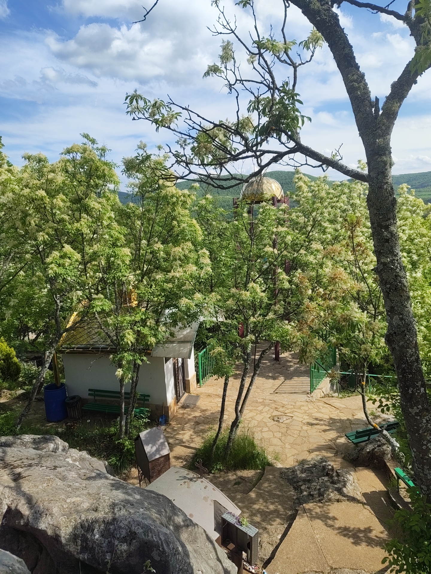 The image depicts an elevated view in Novo Selo, Bulgaria, overlooking a verdant area with trees in bloom. A small, white, one-story building with a golden roof is situated amidst the greenery. A golden dome-shaped structure is partially visible through the trees. A paved walkway or patio with green benches is located near the building. A stone rock formation is visible in the foreground.