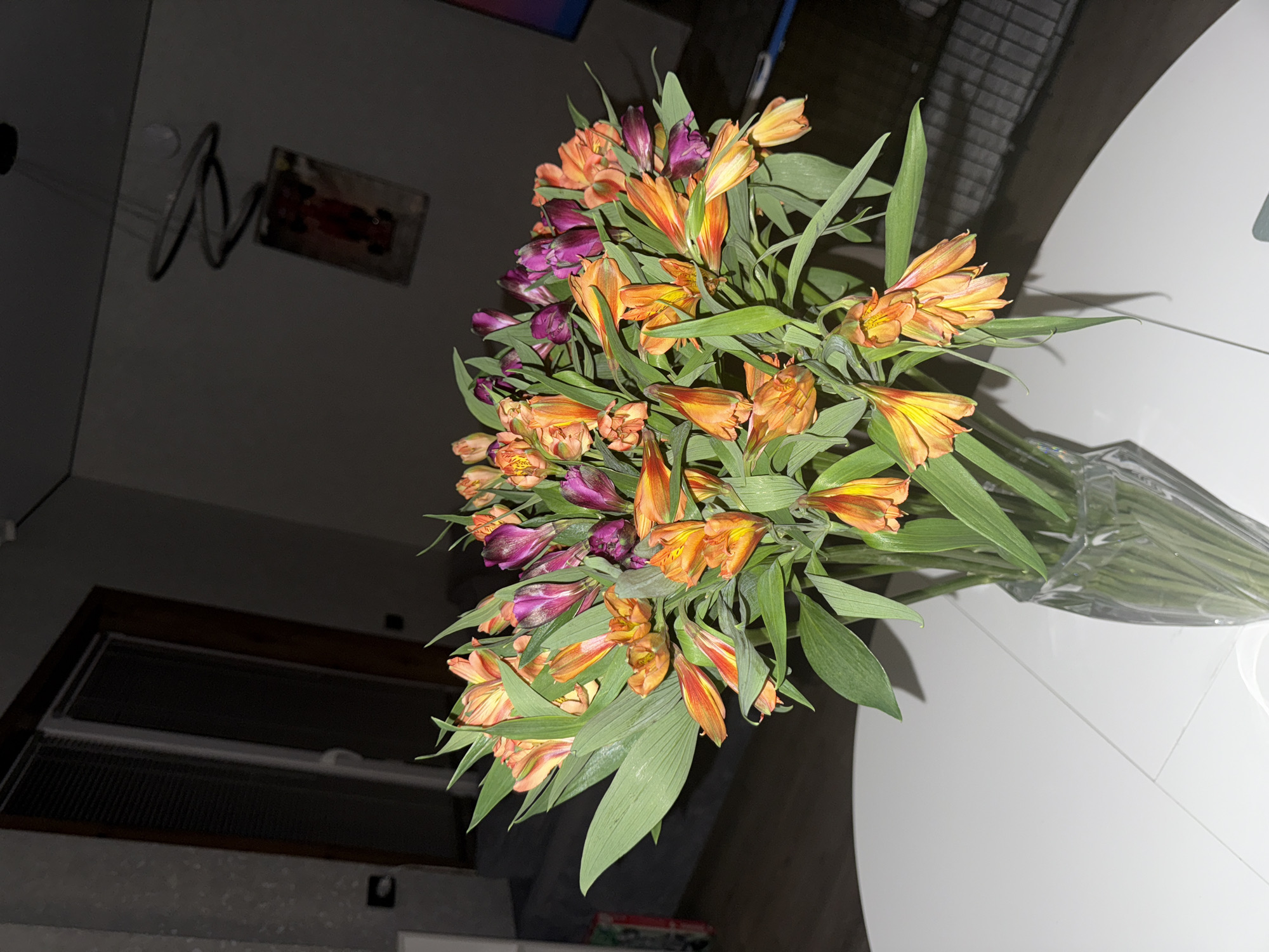 The image shows a bouquet of orange and purple alstroemeria flowers in a clear glass vase, sitting on a white round table. The background is dimly lit, showing a window with blinds and a framed picture on the wall. The location is Лобаново, Russia.