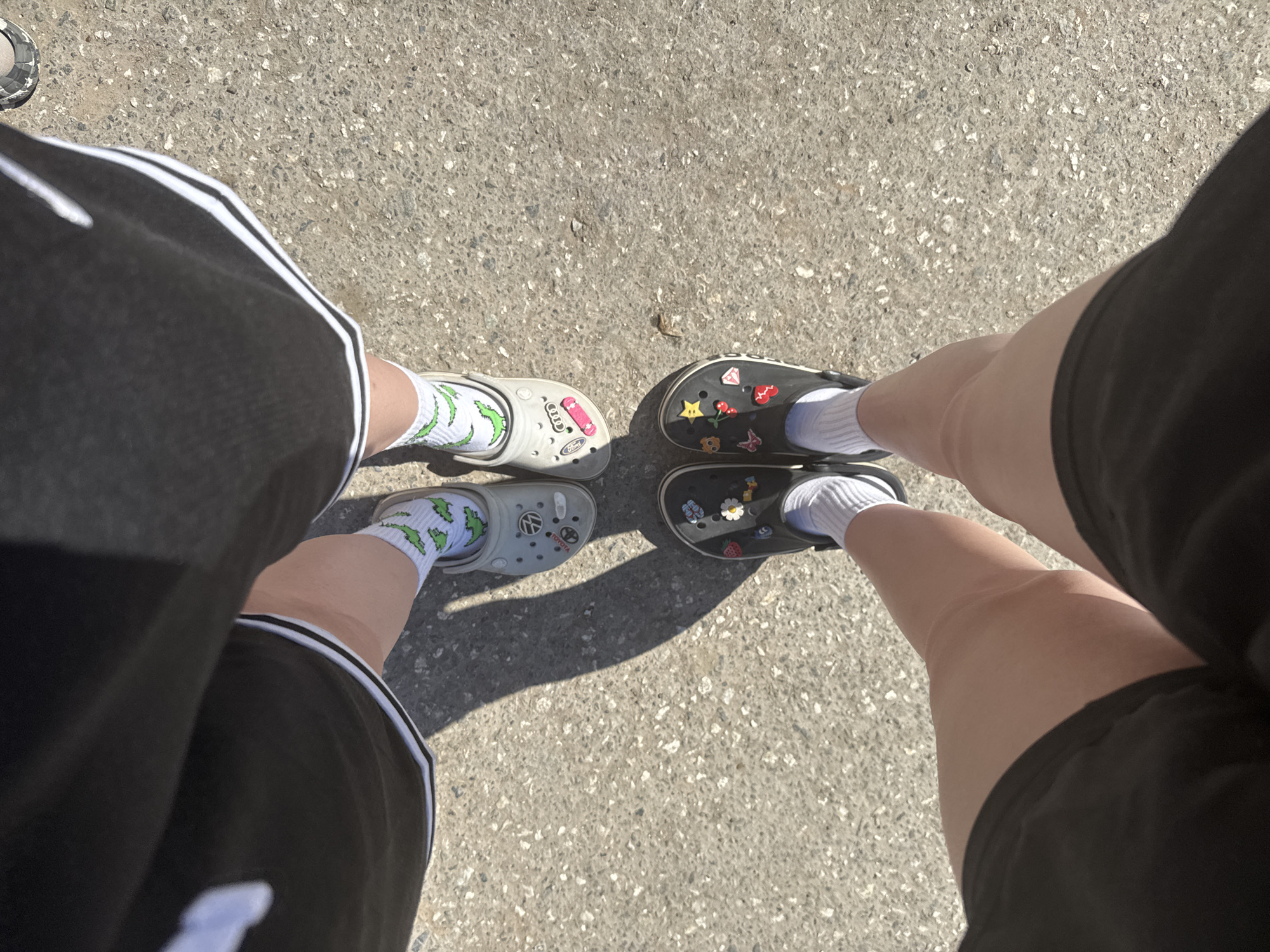 The image is a top-down perspective photograph of two individuals standing on a concrete surface. Both individuals are wearing shorts; the person on the left has on black shorts with white stripes along the sides, while the person on the right has on plain black shorts. The individuals are wearing socks with Crocs. The individual on the left is wearing patterned socks with light-colored Crocs decorated with charms, while the individual on the right is wearing white socks with dark-colored Crocs, which are also adorned with charms. The location is not identifiable as the city of Grishanki, Russia, as there are no distinguishing details to identify that specific location.