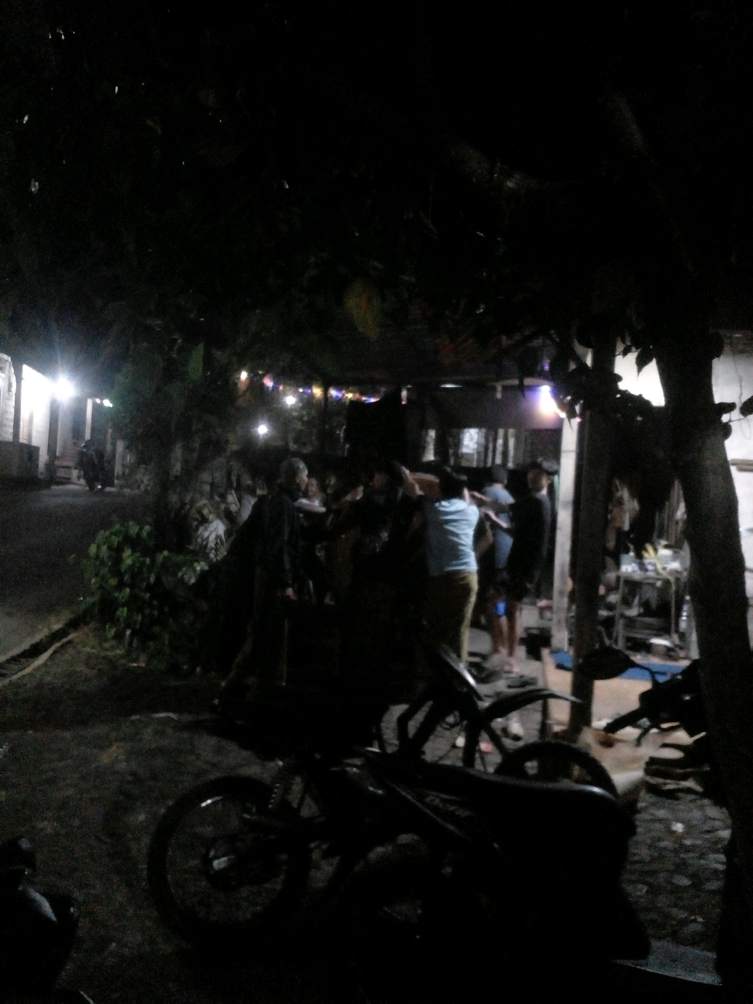 A group of people are gathered outdoors at night, possibly in a residential area of Pungangan, Indonesia. The scene is dimly lit, with artificial lights from buildings and strung decorations providing some illumination. In the foreground, the silhouette of a motorcycle is visible, with its rear wheel prominent. Further back, several individuals are standing and interacting. One person in the middle appears to be wearing a light blue shirt and tan pants. Other figures are less distinct due to the low light conditions. Buildings with illuminated windows are visible on the left side of the frame, and vegetation and trees are present throughout the scene, contributing to a somewhat rustic or tropical atmosphere. The ground appears to be unpaved or covered in dirt and gravel. The overall impression is that of a casual gathering or event taking place in a semi-public, outdoor space after dark.