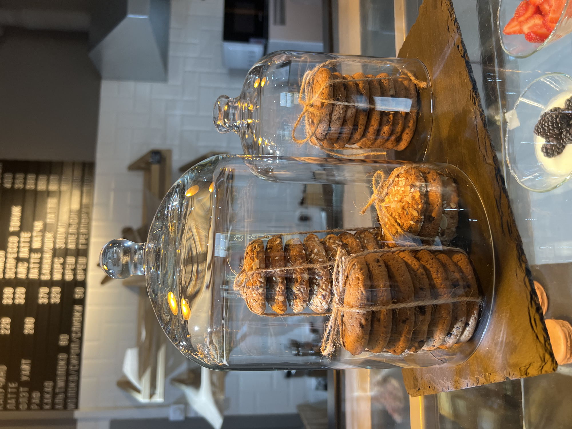 This media file captures a close-up, low-angle shot of a cafe display case, with a focus on two glass cloches filled with cookies. The cookies are stacked neatly and tied with twine. The cloches are placed on a dark, textured surface, possibly slate, which is itself on a wooden board. In the background, a menu board with Cyrillic text is visible, indicating drinks and prices such as "СПРЕССО ТОНИК," "ЛАТТЕ," and "КАКАО." The cafe appears to be well-lit, with warm ambient lighting reflecting off the glass and cookies, suggesting it could be daytime or early evening. To the right of the cookies, a small glass dish holds sliced strawberries, and another contains a white dessert topped with blackberries. The overall scene suggests a bakery or cafe setting, emphasizing fresh baked goods and desserts. There are no people visible in the frame.