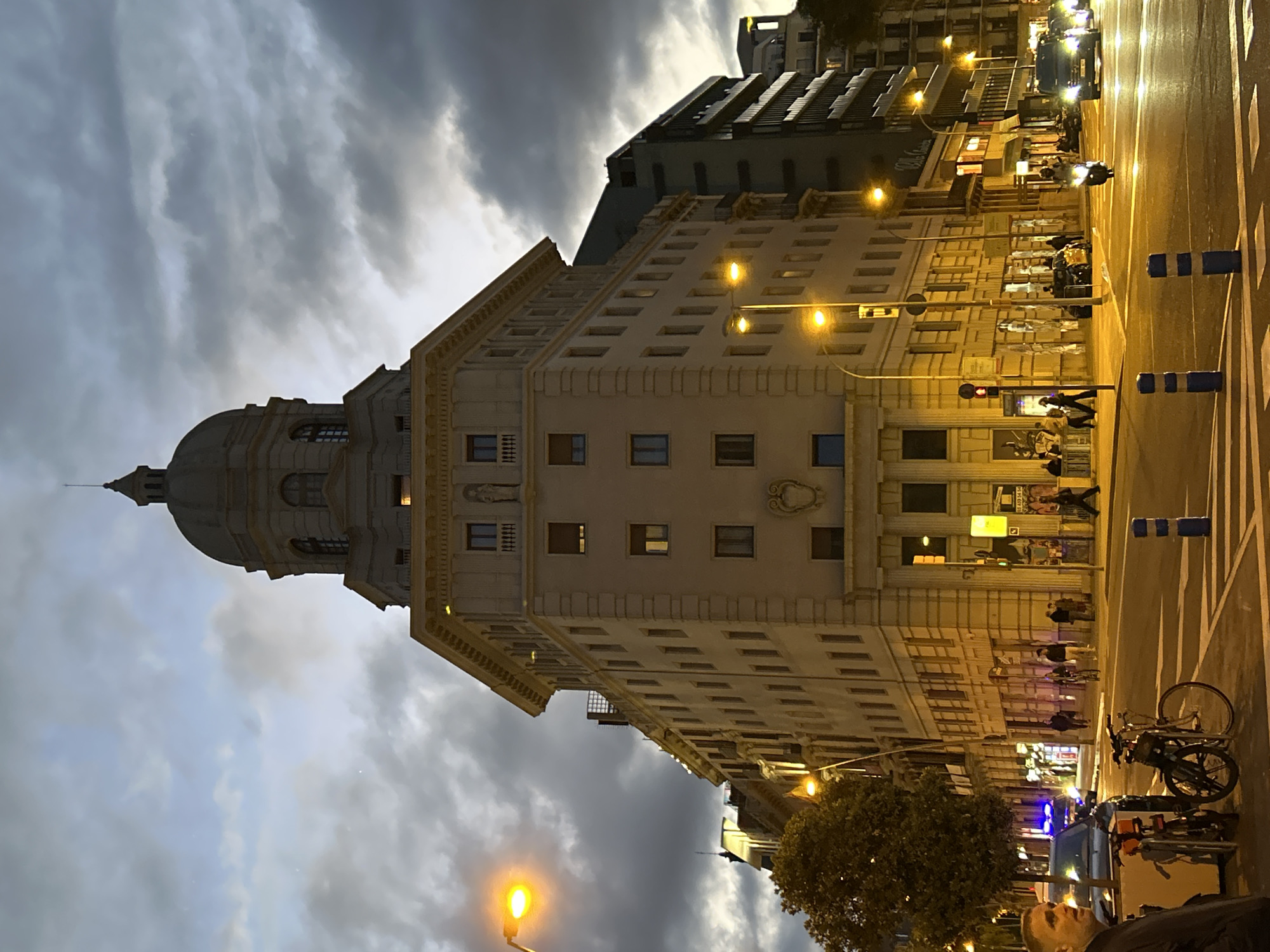The image captures a street scene in Barcelona, Spain at dusk. A prominent, light-colored, multi-story building with a dome-shaped roof and classical architectural details dominates the view. The building has multiple windows and decorative elements.
The street is illuminated by yellow streetlights, creating a warm glow. There are people walking along the sidewalk and crossing the street. Bicycles are parked along the curb. A motor vehicle is present.
The sky is overcast with dark, dramatic clouds. The overall atmosphere is urban and vibrant despite the gloomy sky.