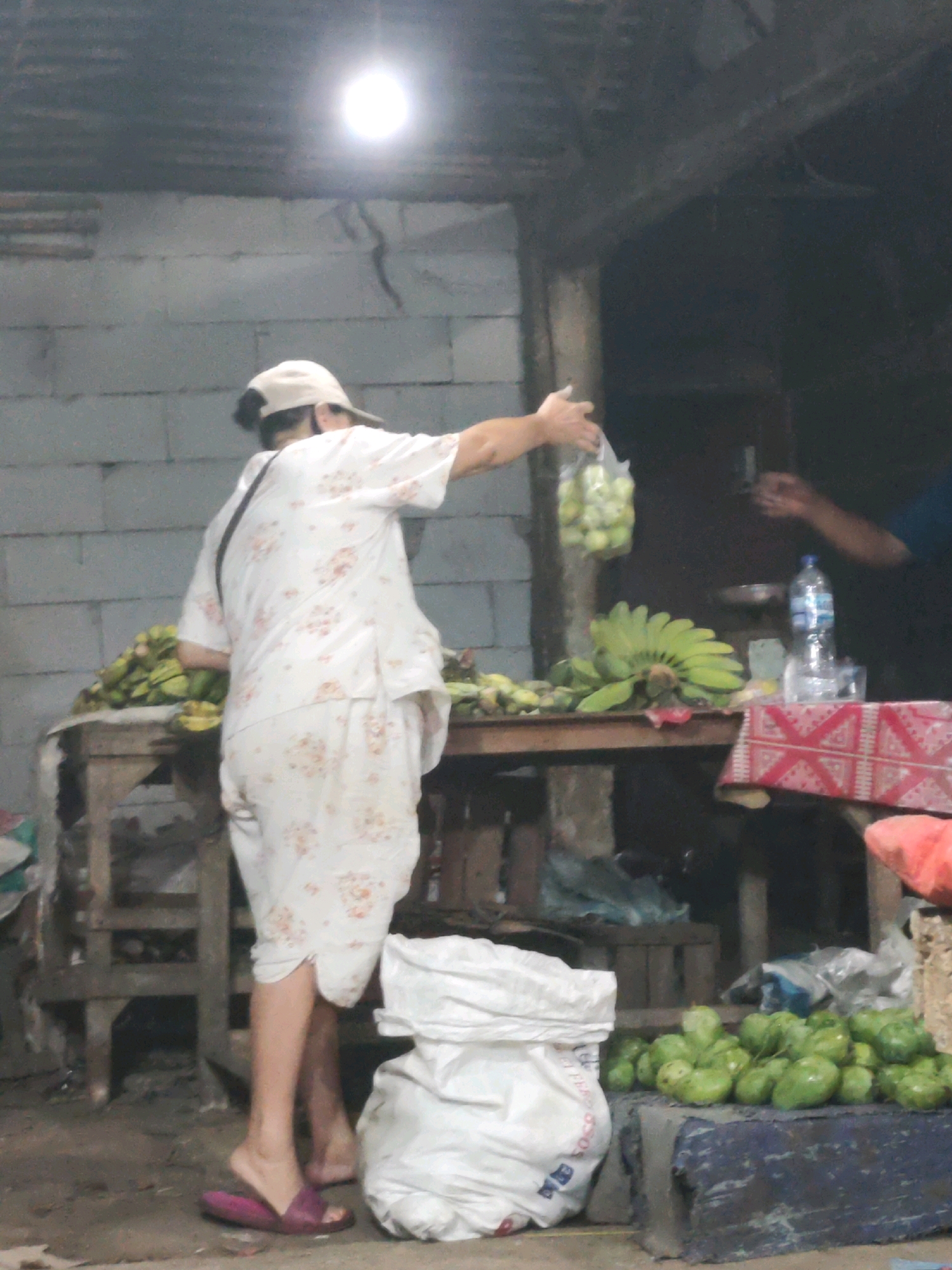 A person wearing a floral patterned white outfit and a cap stands in what appears to be an outdoor market stall in Karawang, Indonesia. The person is holding a plastic bag filled with light green fruits, possibly guavas, and is handing it to another individual whose arm and hand are visible on the right side of the image. In front of the stall, a large pile of green fruits, also appearing to be guavas, is displayed on a blue surface. To the left of the stall, a pile of bananas is visible. A white sack is placed on the ground in the foreground. The background shows a rough, corrugated metal roof and a cinder block wall, suggesting a simple, open-air structure. A bright light bulb hangs from the ceiling, illuminating the scene. The ground is dirt or concrete.
