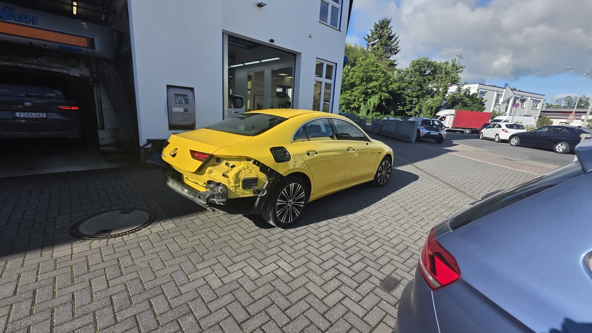 An exterior, eye-level, wide shot shows a bright yellow sedan with significant rear-end damage parked on a brick paver lot next to a white commercial building. The rear bumper and associated components are missing, revealing the car's internal structure. The car has a black fuel door and black rims with silver accents. To the left of the yellow car, a dark-colored SUV is partially visible inside a car wash bay. The building has a large open garage door and several windows. In the background, a street is visible with several other vehicles, including a white sedan, a dark sedan, and a red-and-white truck. Trees and a cloudy sky are also present. The lighting suggests it is daytime.