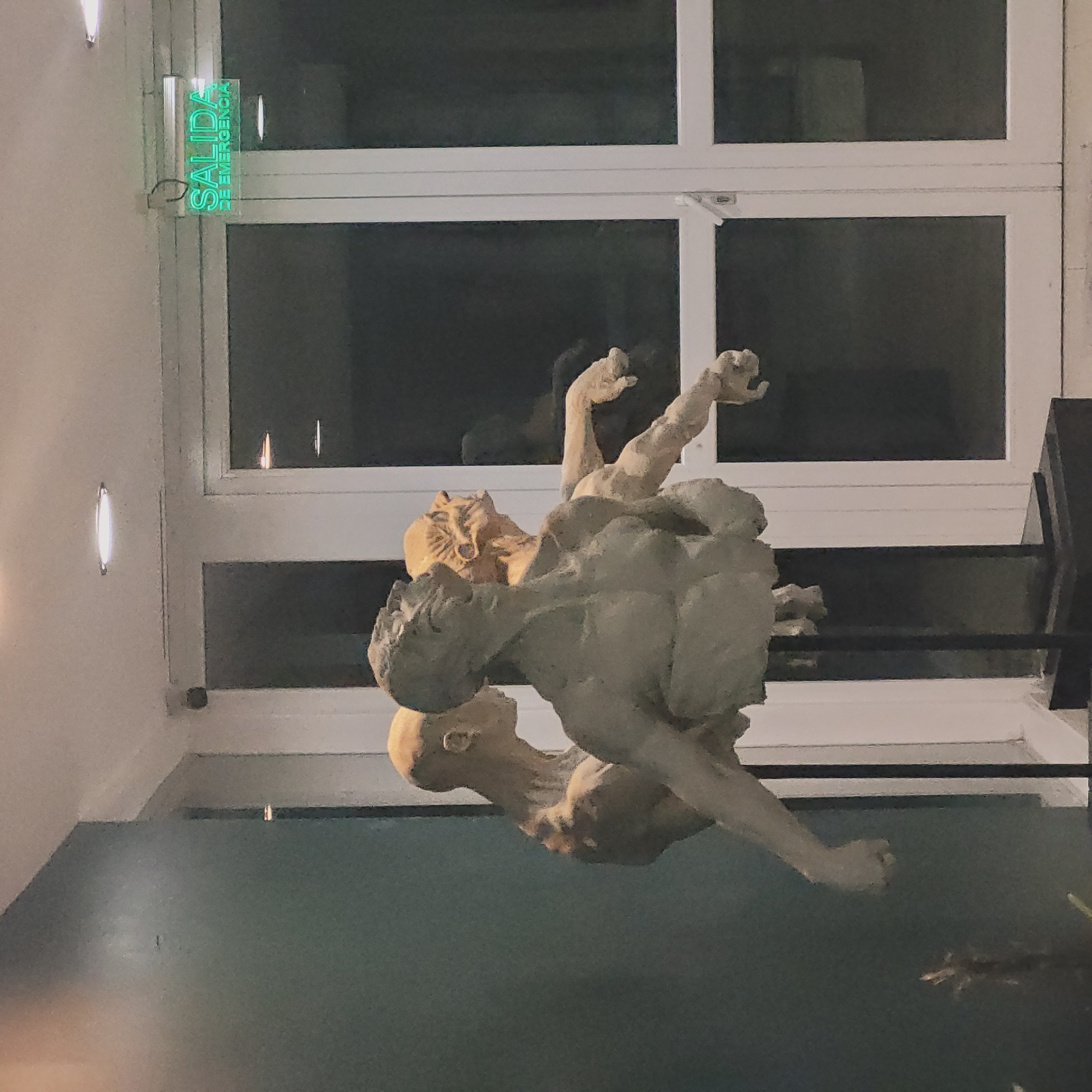 A photograph shows a sculpture in front of a window. The sculpture depicts three nude human figures intertwined in a struggle. The top figure has a bestial or grotesque face, and the sculpture is off-white. The window behind the sculpture has a white frame and is divided into multiple panes. Above the window, a green illuminated sign reads "SALIDA DE EMERGENCIA" (Emergency Exit). The wall is light beige, with two oval-shaped lights on the left side. The photograph appears to have been taken indoors, looking up at the sculpture. The bottom part of the image shows a dark surface, likely a wall or floor. Given the context of "San Fernando del Valle de Catamarca, Argentina", this image likely depicts a sculpture in a building within that city, with the "SALIDA DE EMERGENCIA" sign indicating an emergency exit.