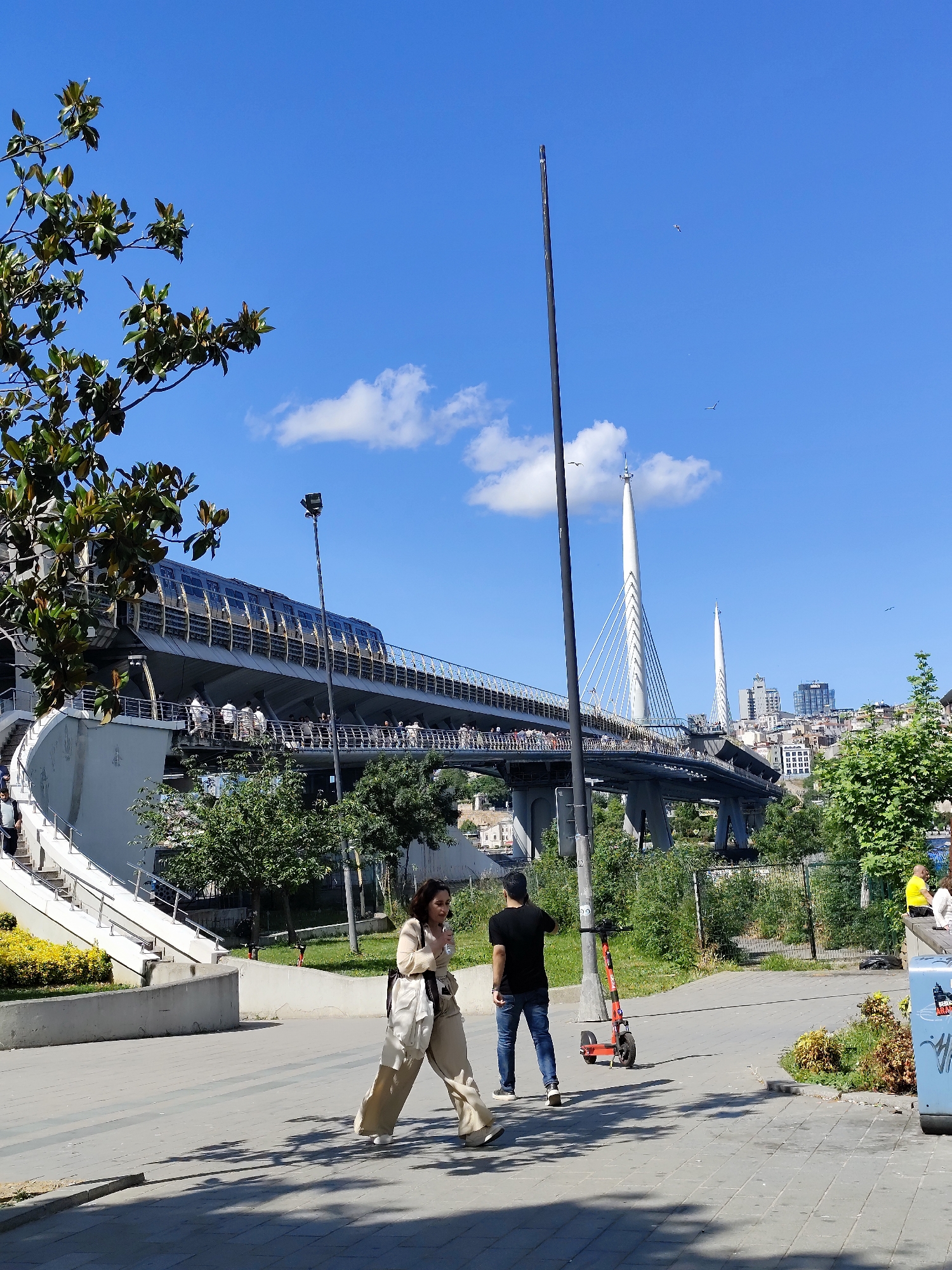 The image shows an outdoor scene in a city, possibly Istanbul, based on the bridge architecture in the background. A woman in a tan outfit walks to the left, while a man in a black shirt and jeans stands to the right next to a red scooter. In the background, a modern bridge carries a train or tramway, crowded with people. The bridge's structure includes two tall, white pylons. In the far background, a cityscape is visible. The weather appears sunny with a few clouds in the blue sky. The surrounding area includes greenery, trees, and walking paths.