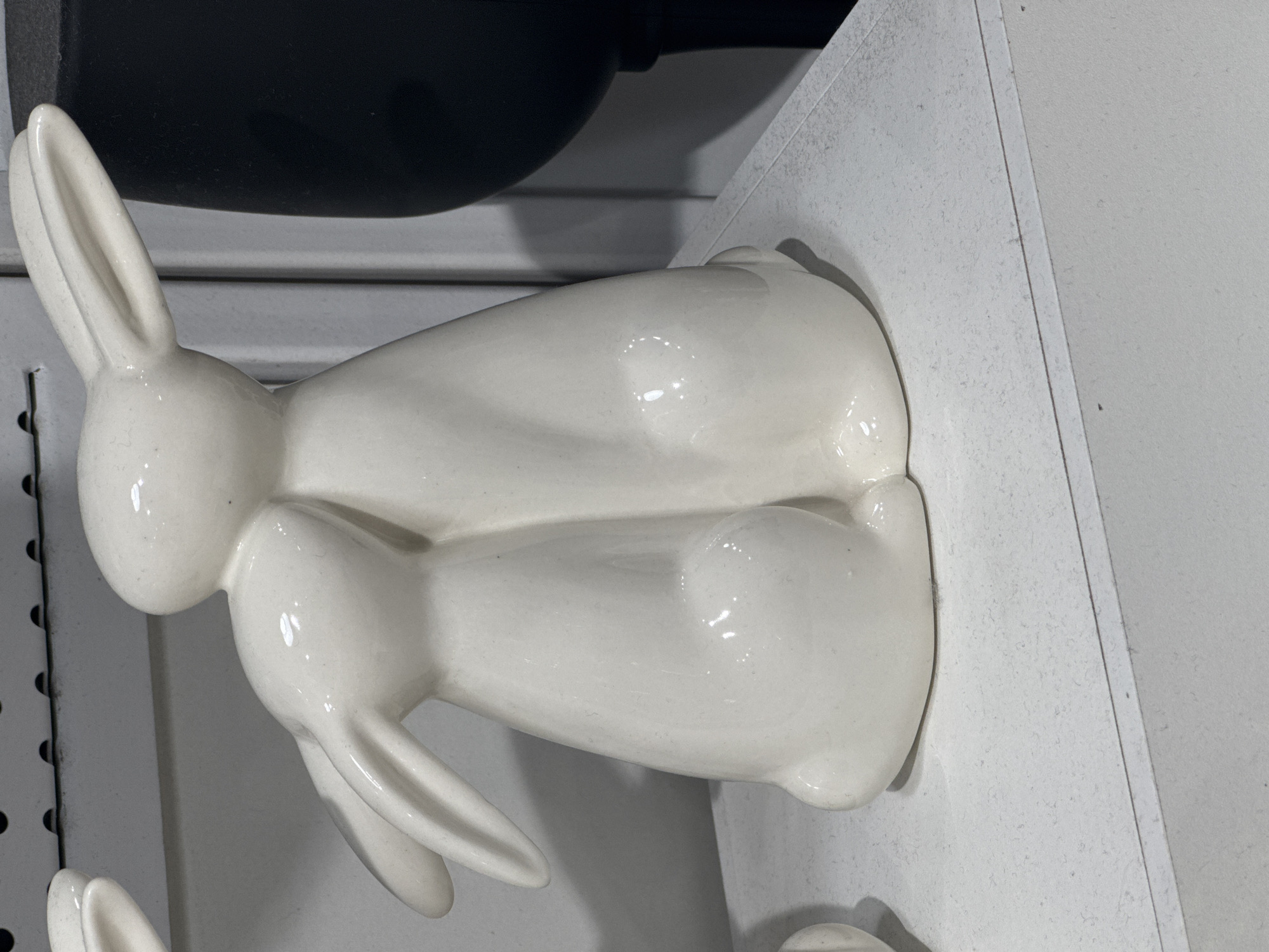 A close-up shot focuses on a pair of white ceramic bunny figurines embracing each other. The bunnies are positioned on a white shelf. The larger bunny is behind and slightly above the smaller bunny, with its head resting on the smaller bunny's head. Both bunnies have long, upright ears. The figurines appear to be smooth and glossy, reflecting the light. In the background, a portion of a large black vase is visible on the right, and a white wall with some shelving detail is on the left. The image has a shallow depth of field, blurring the background elements and keeping the bunnies in sharp focus. The scene suggests a decorative display, possibly in a home or retail environment, and is not identifiable as being in Ufa, Russia.