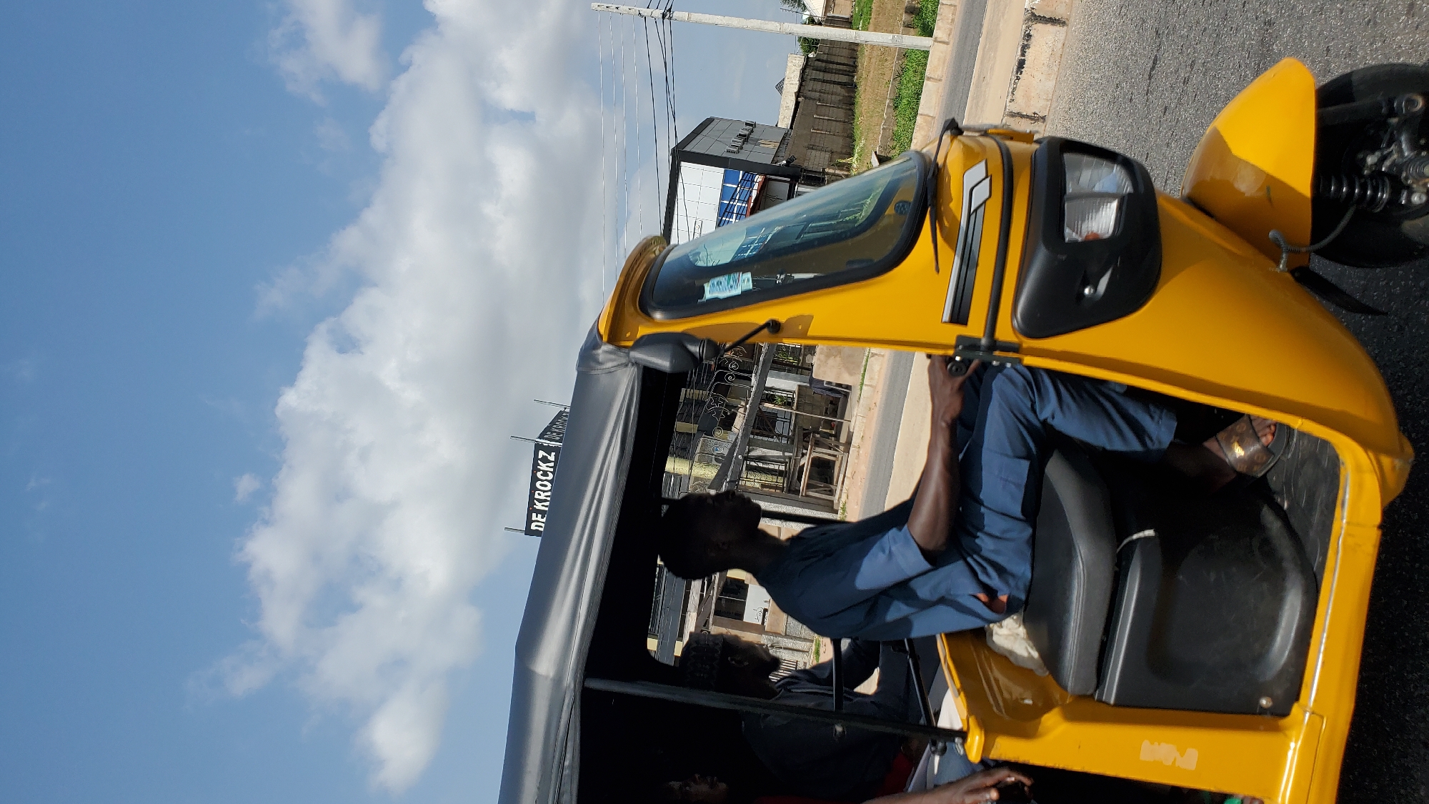 The media file shows a side view of a yellow auto-rickshaw with two people inside. The driver, wearing a blue uniform, is visible in the front seat. Another person is seated in the back. The scene appears to be set during the day, with a bright blue sky and white clouds. The background shows buildings, a road, and some vegetation, suggesting an urban or semi-urban environment in Jalingo, Nigeria. A sign with the text "DE KROCKZ" is visible on the left side of the image. The overall impression is of a typical day of transportation in a Nigerian city.