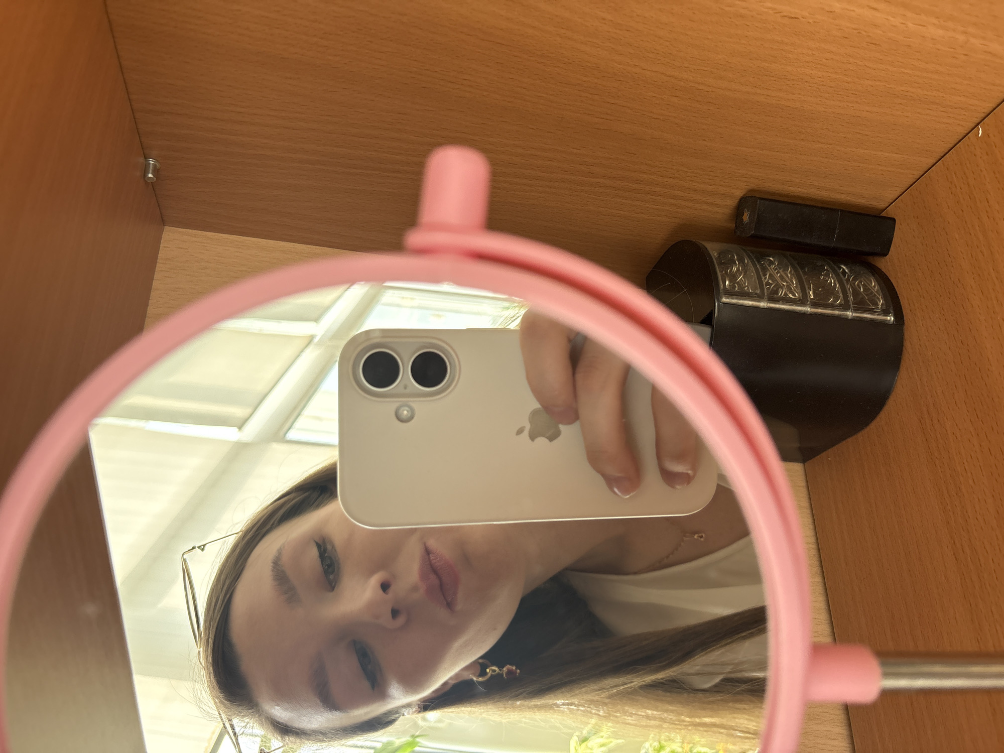 A woman in Izhevsk, Russia, is taking a selfie in a pink, circular mirror. She is holding a white iPhone with two cameras. The mirror is positioned inside a light brown wooden cabinet or shelf. On the top right of the shelf, there is a small, black rectangular object next to a black, decorative object. The woman has long brown hair and is wearing glasses and a white top. Her facial expression is neutral.