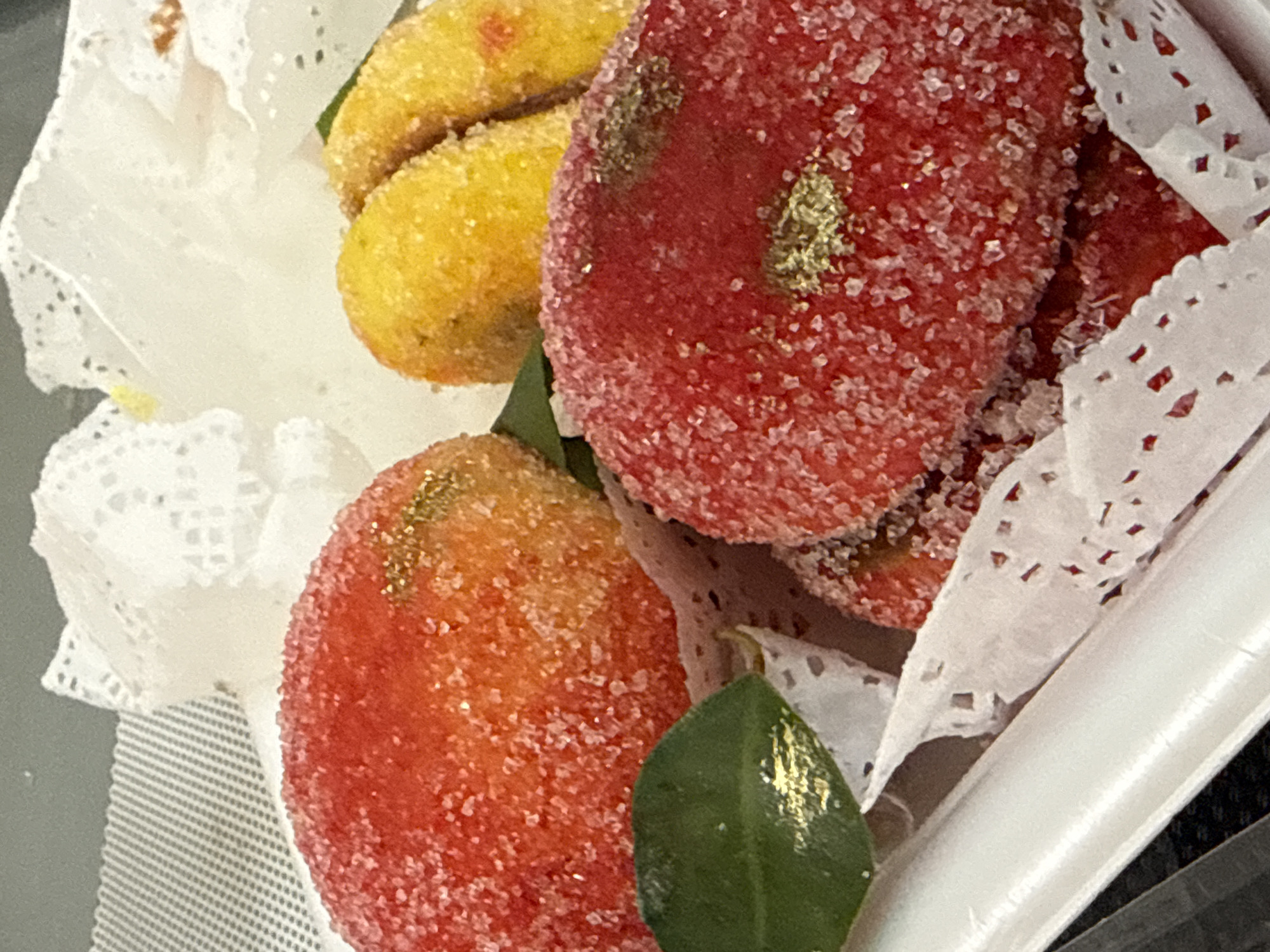 The image shows a close-up of a white container filled with several pastries. The pastries are round and oval-shaped, coated in sugar crystals and some appear to have gold leaf accents. There are red and yellow colored pastries, resembling peaches and bananas in shape. Some of the pastries are nestled in decorative white paper. A green leaf is also visible on one of the red pastries. The background is out of focus and appears to be a grey surface with a patterned texture. The lighting is bright, highlighting the sugar coating and gold leaf on the pastries.