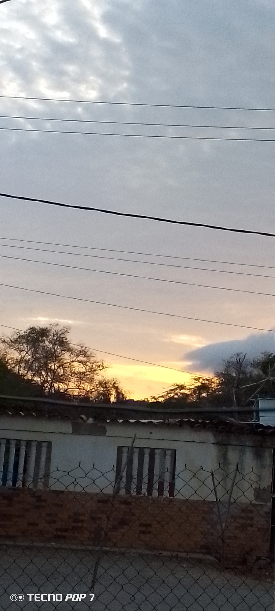 The image shows a view towards the horizon in Altagracia, Venezuela, featuring a sunset or sunrise. In the foreground is a fence, behind which a building with windows can be seen. Above the building are silhouettes of trees. Power lines stretch across the upper part of the frame against a cloudy sky with a hint of orange light on the horizon. The "Tecno Pop 7" watermark is visible in the lower left corner.
