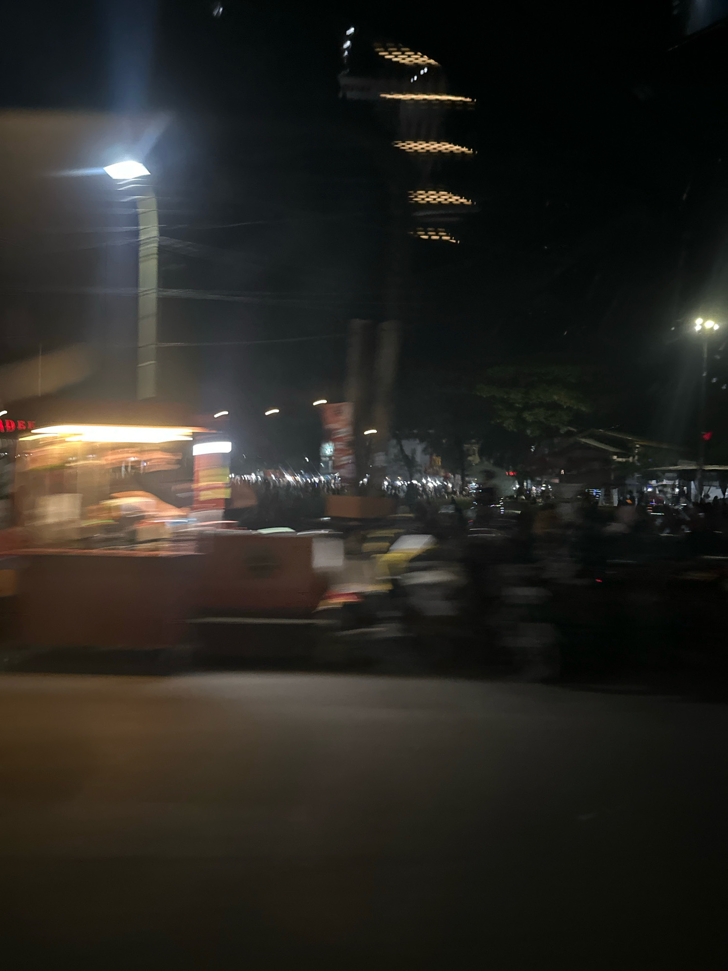 The image depicts a blurred nighttime scene in the City of Binjai, Indonesia, likely from a moving vehicle. In the foreground, a food stall or cart is visible, illuminated by bright lights, suggesting it is open for business. Behind the stall, the environment appears to be a street or public area with multiple individuals, possibly customers or passersby, also blurred due to motion. Various lights from street lamps, signs, and possibly building interiors are present, creating a dynamic and somewhat chaotic urban atmosphere. Trees and other structures are discernible in the background, contributing to the impression of a busy city at night. The overall impression is one of urban activity, captured in a transient moment.