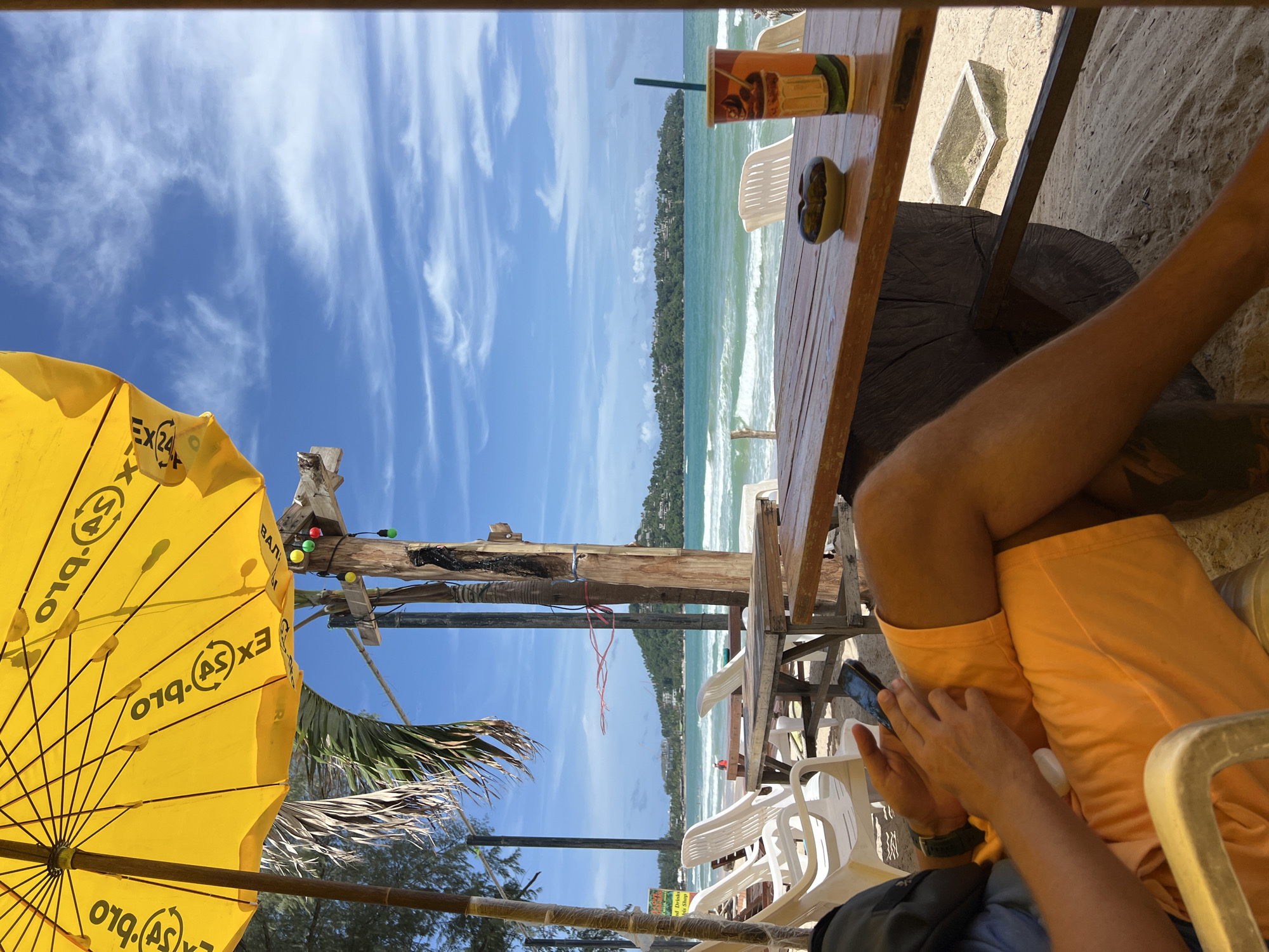 A person sits at a wooden table on a sandy beach, holding a smartphone. The person is wearing orange shorts. In front of them on the table is a drink in a cup and a small ceramic ashtray. Behind the person, white plastic chairs are visible, and beyond that, the ocean with waves and a distant tree-covered island under a blue sky with scattered clouds. A yellow umbrella shades part of the scene. String lights are attached to a wooden pole in the background. The location is a beachside establishment.