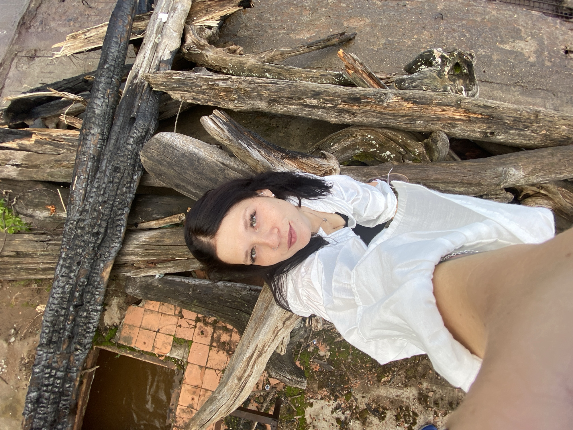 A high-angle, eye-level medium shot captures a young woman with dark hair and light skin, adorned with subtle makeup and a nose ring, standing amidst a pile of weathered and burnt wood. She wears a white, unbuttoned button-down shirt over a black top. Her left arm extends towards the camera, obscuring part of her lower torso, while a silver necklace is visible around her neck. The background is an outdoor scene, with elements of an abandoned or dilapidated area, possibly a construction site or junkyard. The ground is a mix of dirt, moss, and possibly debris. To the left of the woman, a shallow pool of murky, brown water is visible, bordered by cracked orange tiles. The overall impression is one of decay and abandonment.