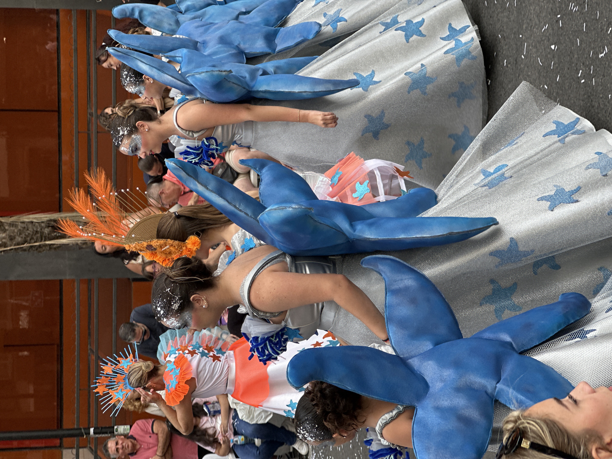 The image captures a vibrant outdoor scene featuring multiple individuals in elaborate costumes, consistent with a festive event or parade in the city of Unknown, Unknown. In the foreground and midground, several women are prominently displayed in matching themed attire. The primary costume consists of a shimmering silver sleeveless dress with a full, floor-length skirt. The skirt is adorned with numerous light blue starfish patterns. Attached to the backs and sides of these dresses are large, soft, sculptural elements crafted from a vibrant blue fabric, resembling stylized wings or aquatic appendages. The women wearing these costumes have elaborate makeup, including silver glitter on their foreheads and temples, and intricate patterns of silver and blue face paint. Their hair is styled in updos, with some also featuring glitter. Specifically, from left to right among the main subjects: * A partial view of a young woman's face in the extreme lower-left corner, with blonde hair and sunglasses perched on her head. * A woman in the silver and blue costume, seen from the side, with her right arm visible, wearing a delicate gold bracelet. * Behind her, another woman in a similar costume, her face partially visible with the glitter makeup. * Further right, a third woman in an identical costume, seen mostly from the back, displaying the blue fabric elements. * Behind and to the left of the silver-clad women, another participant is visible, wearing a white top with an orange ruffled collar and a large, intricate headpiece featuring spiky orange and blue elements with stars. Her white skirt also has orange sections and blue starfish. She is shown with her hands near her headpiece, seemingly adjusting it. * To the far right, part of another woman in an orange feathered headpiece can be seen. In the background, behind the costumed individuals, a diverse crowd of onlookers is present. Notable figures include a man with short grey hair and glasses, wearing a pink polo shirt, with his arms crossed, observing the scene from the left. Another man in a dark shirt and sunglasses is visible further back. Parts of other adults and children can be discerned, some seated. The immediate backdrop consists of a large building with a textured, reddish-brown or terracotta facade featuring horizontal metal slats or railings across its stories. The ground beneath the participants is a dark gray pavement, scattered with small white specks, possibly confetti or debris.