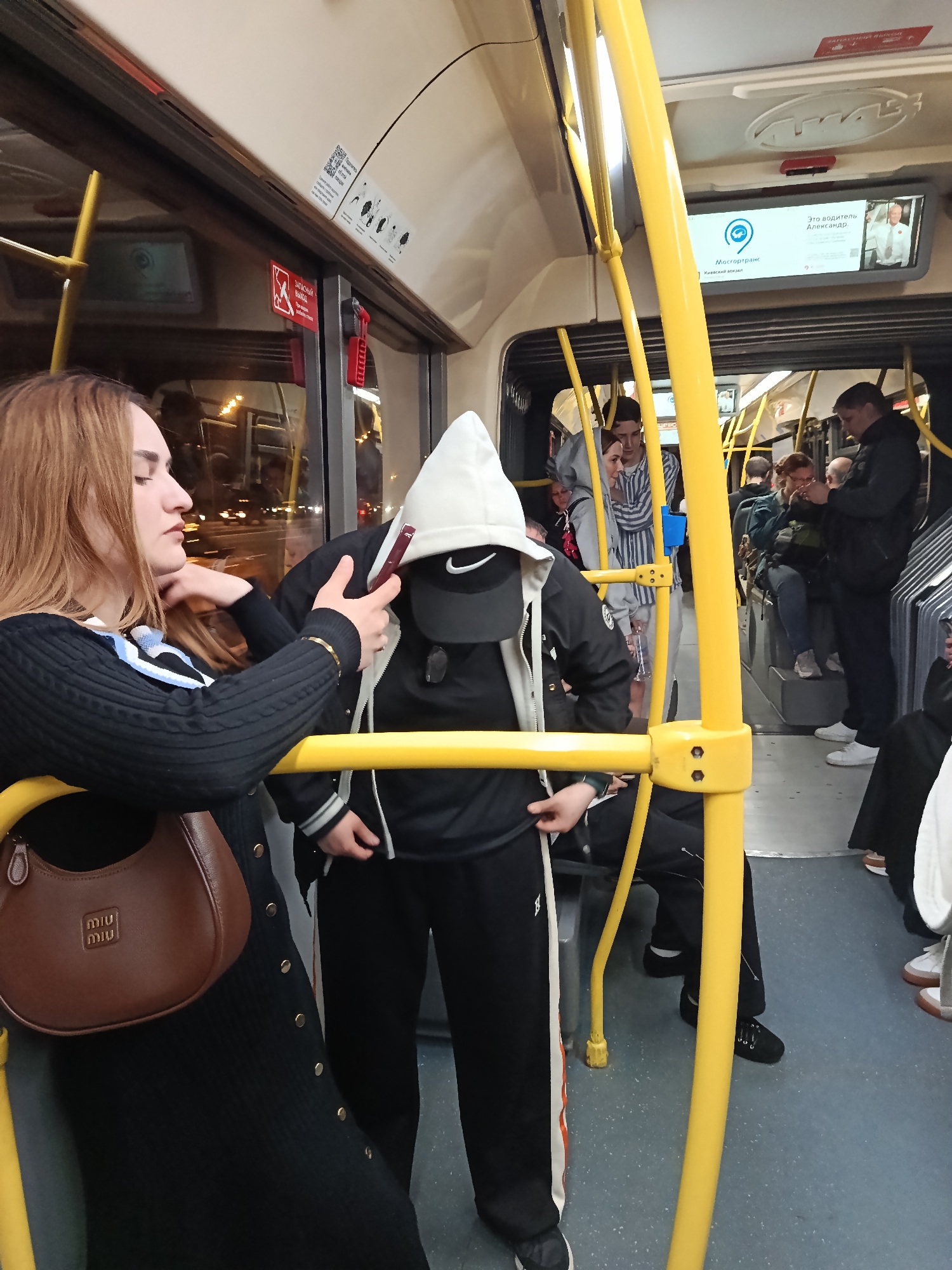 The image depicts a scene inside a bus, likely a public transportation vehicle in an urban setting.
A woman with long, reddish-brown hair is on the left side of the frame. She is wearing a dark, possibly black, button-up dress or sweater, and a brown handbag with a "MIU MIU" logo is slung over her shoulder. She is holding a red cellphone to her ear, seemingly engaged in a phone call.
In front of her stands a person dressed in a black Nike baseball cap, a hooded sweatshirt, and black sweatpants with white and orange stripes down the sides. The person has pulled the hood of the sweatshirt up, obscuring their face.
Behind the two, several other passengers are visible. Some are standing, others are seated.
The bus interior is characterized by yellow handrails. A digital display above shows the route information, likely a city transportation system. The environment suggests an urban setting at night, as indicated by the reflections on the bus windows.