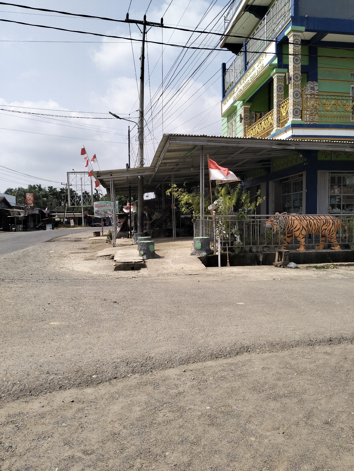 In Serdang Jaya, Indonesia, a two-story building with blue and green accents stands beside a covered walkway. Indonesian flags fly from the walkway's support poles. In front of the building, a tiger statue is displayed, and a metal fence separates it from the road. The road itself is paved with asphalt and gravel, showing signs of wear and tear. The area is lined with trees and utility poles carrying numerous wires. Other buildings and structures are visible in the background, suggesting a mixed-use or commercial area. The sky is partly cloudy.