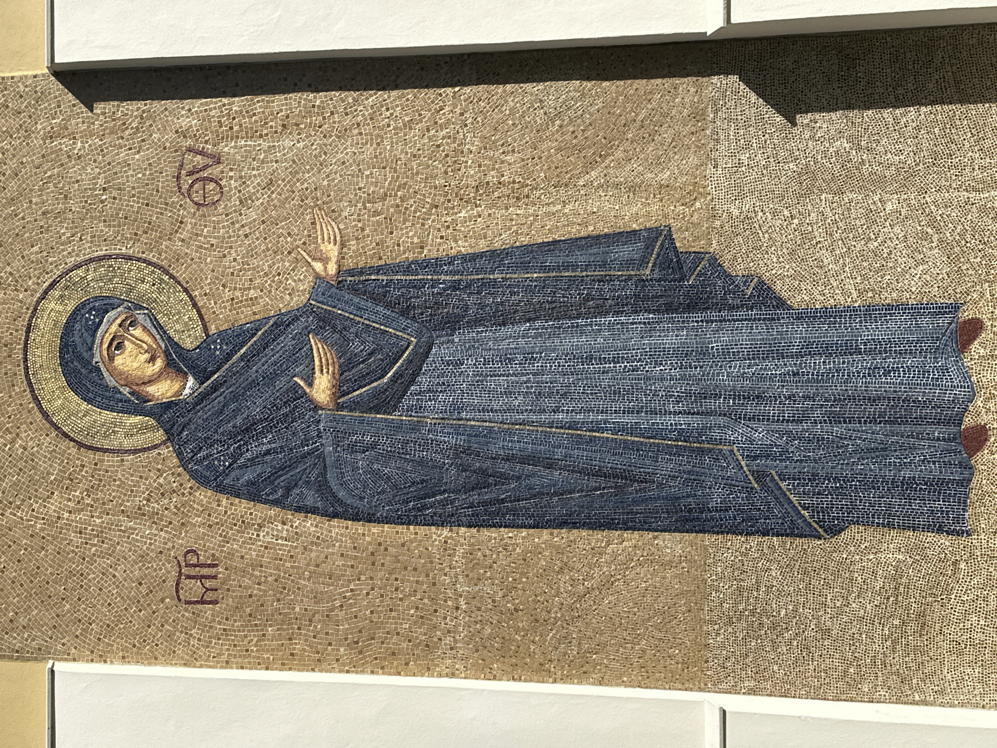 A full-length mosaic depicting the Virgin Mary is presented on an exterior wall in Maldon, United Kingdom. The figure, rendered in an iconographic style using numerous small tesserae, wears a dark blue maphorion with gold accents, covering her head and body. A golden halo encircles her head. Her face is pale with a solemn expression, and her eyes are cast downward. Both hands are outstretched forward, palms open and upward in a gesture of reception. To the left of her head, the red Greek monogram 'MP' (Μήτηρ) is visible; to the right, the red Greek monogram 'ΘV' (Θεοῦ) identifies her as 'Mother of God'. The mosaic features a golden-brown background and is bordered vertically by white architectural elements. A distinct shadow from the right border falls across a portion of the mosaic, suggesting a directed light source.
