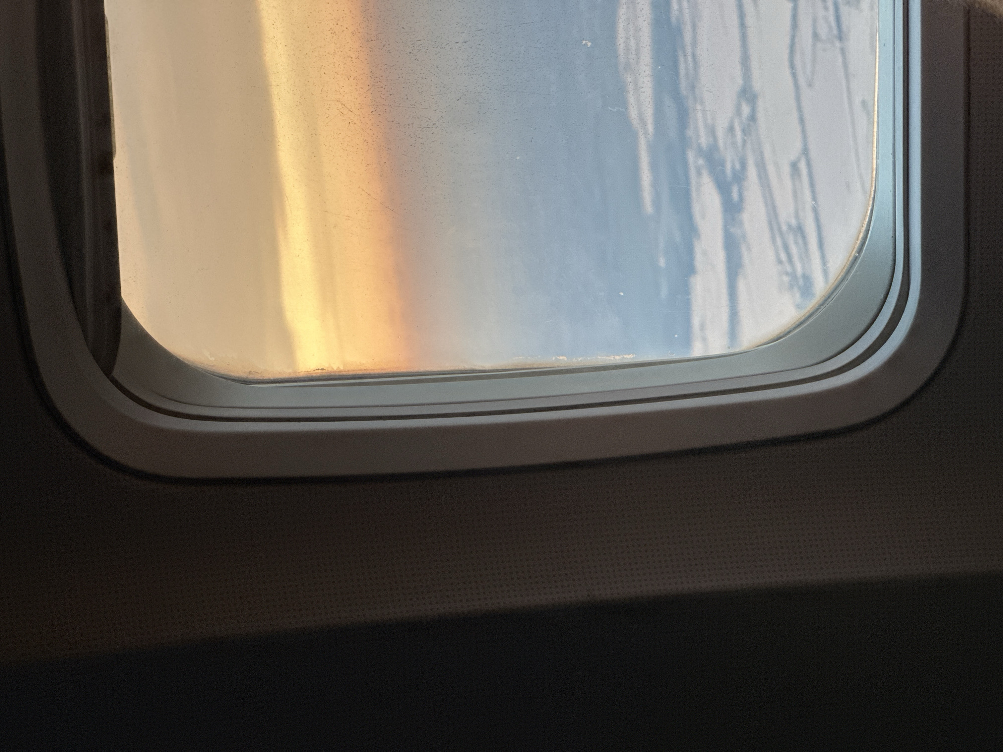 The image is taken from inside an airplane, looking out of a window. The window frame is a light gray color and shows signs of wear. The view outside the window is of clouds and a sky tinged with the warm colors of sunset or sunrise, possibly indicating a flight at dawn or dusk. The clouds below are white and spread out, suggesting a view from a high altitude. The interior of the airplane's cabin is visible on the left side of the frame, with a dark textured wall. There are no people or specific landmarks visible in the image. The location in the city of Tzafria, Israel, cannot be determined from the visual information provided in the image.