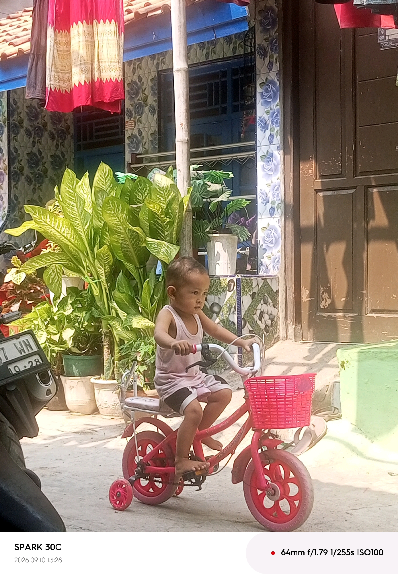 A young child, wearing a light pink tank top and shorts with black trim, sits on a red children's bicycle with training wheels. The child's feet are on the pedals and their hands are on the handlebars, appearing to be in motion. The bicycle has a red basket attached to the front. In the background, there are several potted plants, one large with broad green leaves. Behind the plants, a building is visible with blue trim and patterned tiles featuring blue roses. A wooden door is also present to the right of the building. To the left of the child, part of a motorcycle with a license plate reading "37 WN 10.28" is visible. The scene appears to be outdoors on a paved or dirt surface. The lighting suggests it is daytime, with shadows cast on the ground. The image metadata indicates the photograph was taken with a SPARK 30C camera on September 10, 2026, at 13:28, with settings of 64mm, f/1.79, 1/255s, and ISO 100. This scene is set in Tegal, Indonesia.