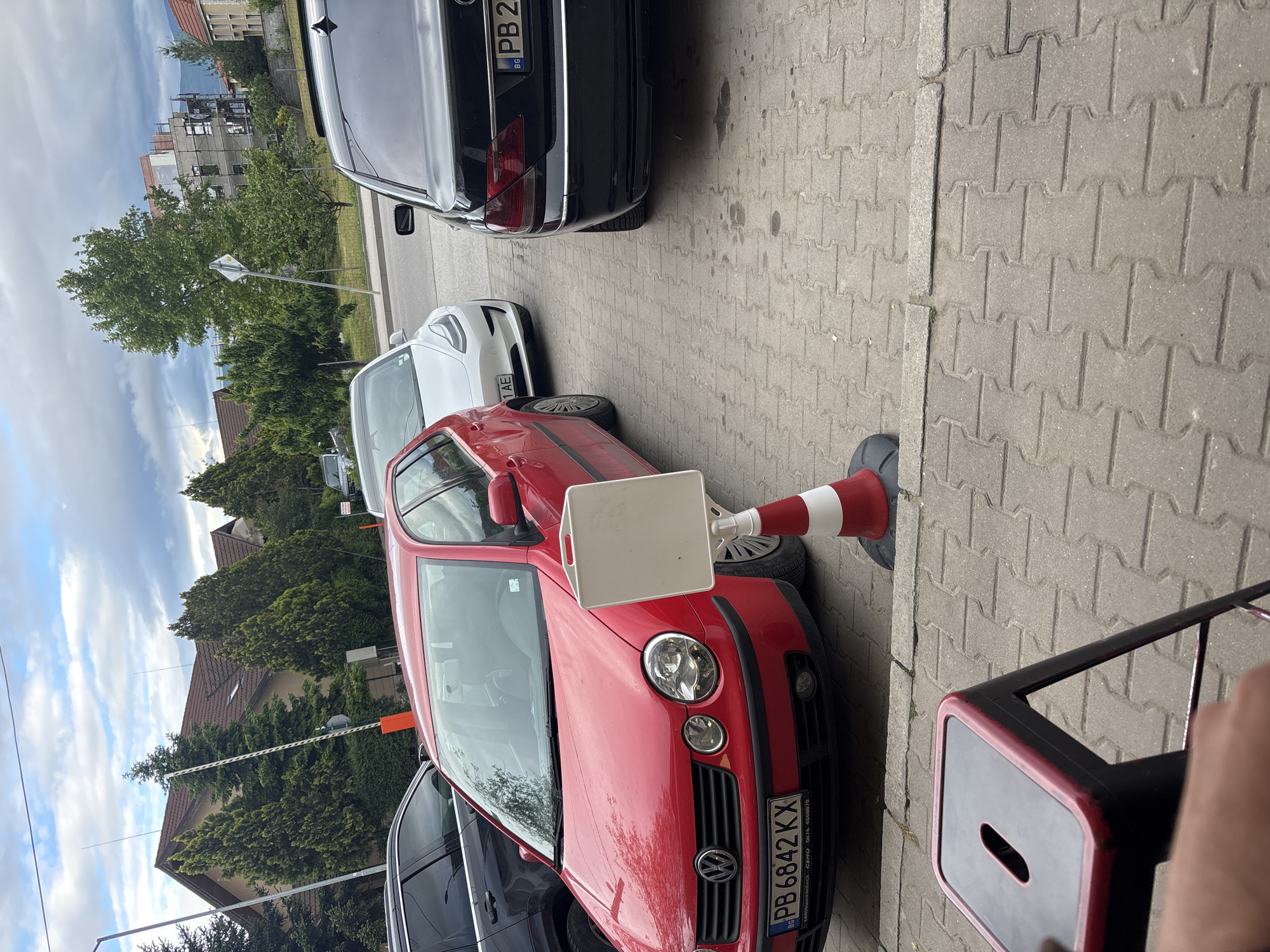 The image depicts a street scene in Plovdiv, Bulgaria. A red Volkswagen Polo, registration number PB 6842 KX, is parked on a paved surface. A traffic cone with white and red stripes rests against the car's front left side. Adjacent to the Polo are other parked cars, including a white vehicle and a black vehicle with a registration number starting with "PB 2". A black stool with a red trim is situated near the Polo on the right side of the frame. Buildings and trees are visible in the background, beneath a cloudy sky.