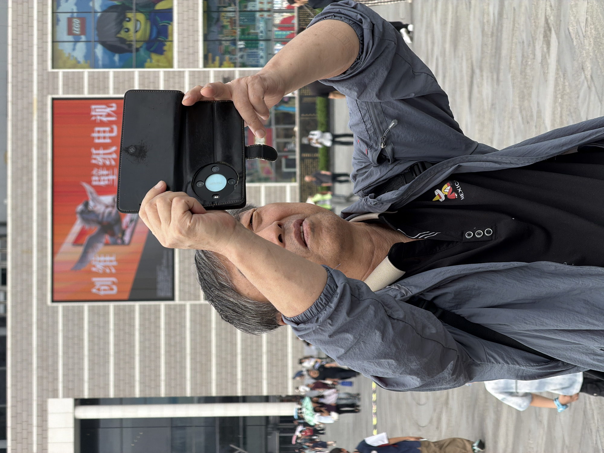 A middle-aged East Asian man with short, graying hair is holding a black phone case with a phone inside in both hands, taking a picture outdoors. He is wearing a gray zipped jacket over a black polo shirt with a graphic of a person in a sombrero and the word "MEXICO" on it. The man's face is turned slightly upwards and to the right, with his eyes looking through the phone. In the background, there is a large building with a brick facade and horizontal white stripes. A large advertisement with Chinese characters and an image of a bird is displayed on the building. To the right of this ad, there is another large window or display featuring a Lego character. Several other people, blurred due to motion, are visible in the distance, some holding umbrellas, suggesting a possibly overcast day. The ground appears to be paved. The scene is set in the Yuzhong District of Chongqing, China.