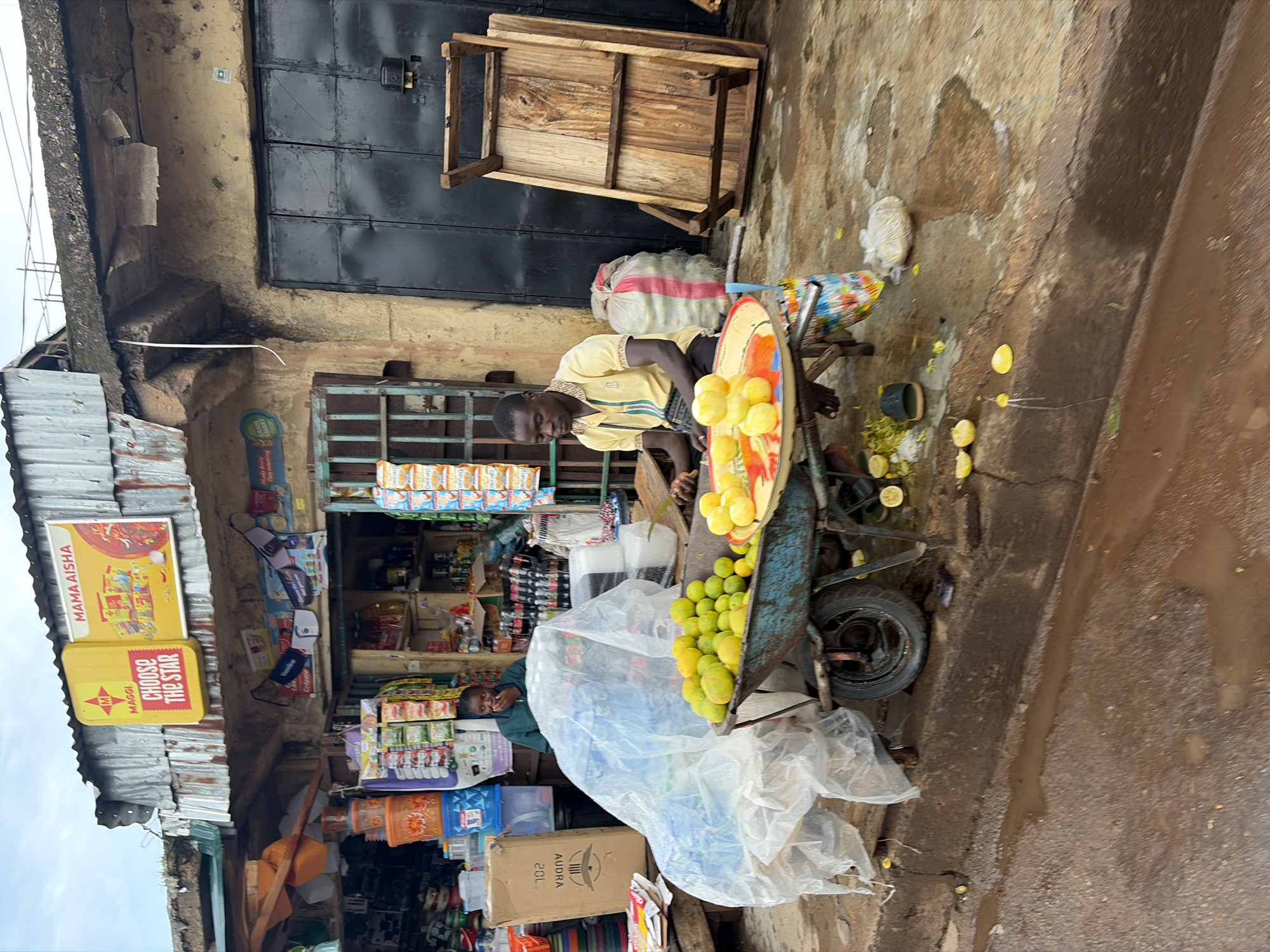 This is a photograph of a street vendor in Jos, Nigeria, likely operating from a small shopfront. The vendor, a man wearing a yellow collared shirt and dark trousers, is seated and appears to be preparing fruit, possibly oranges or lemons, which are displayed in a metal tray in front of him. He is positioned next to a blue wheelbarrow filled with more fruit, covered partially by a translucent plastic sheet. Scattered around the ground are pieces of cut fruit and what appear to be peelings.
The shopfront itself shows signs of wear and tear, with a corrugated metal roof and a weathered concrete facade. Above the shop, signage is visible, including advertisements for "MAGGI CHOOSE THE STAR" and "MAMA AISHA". The shop appears to sell various goods, with shelves stocked with packaged items and bottles visible through an open doorway. A large black metal door is visible to the right of the shop. The ground is wet and muddy, suggesting recent rainfall. In the background, another person, possibly a customer, is partially visible inside the shop. A wooden table is propped up against the wall near the vendor. The overall scene depicts a typical bustling market environment in Jos.
