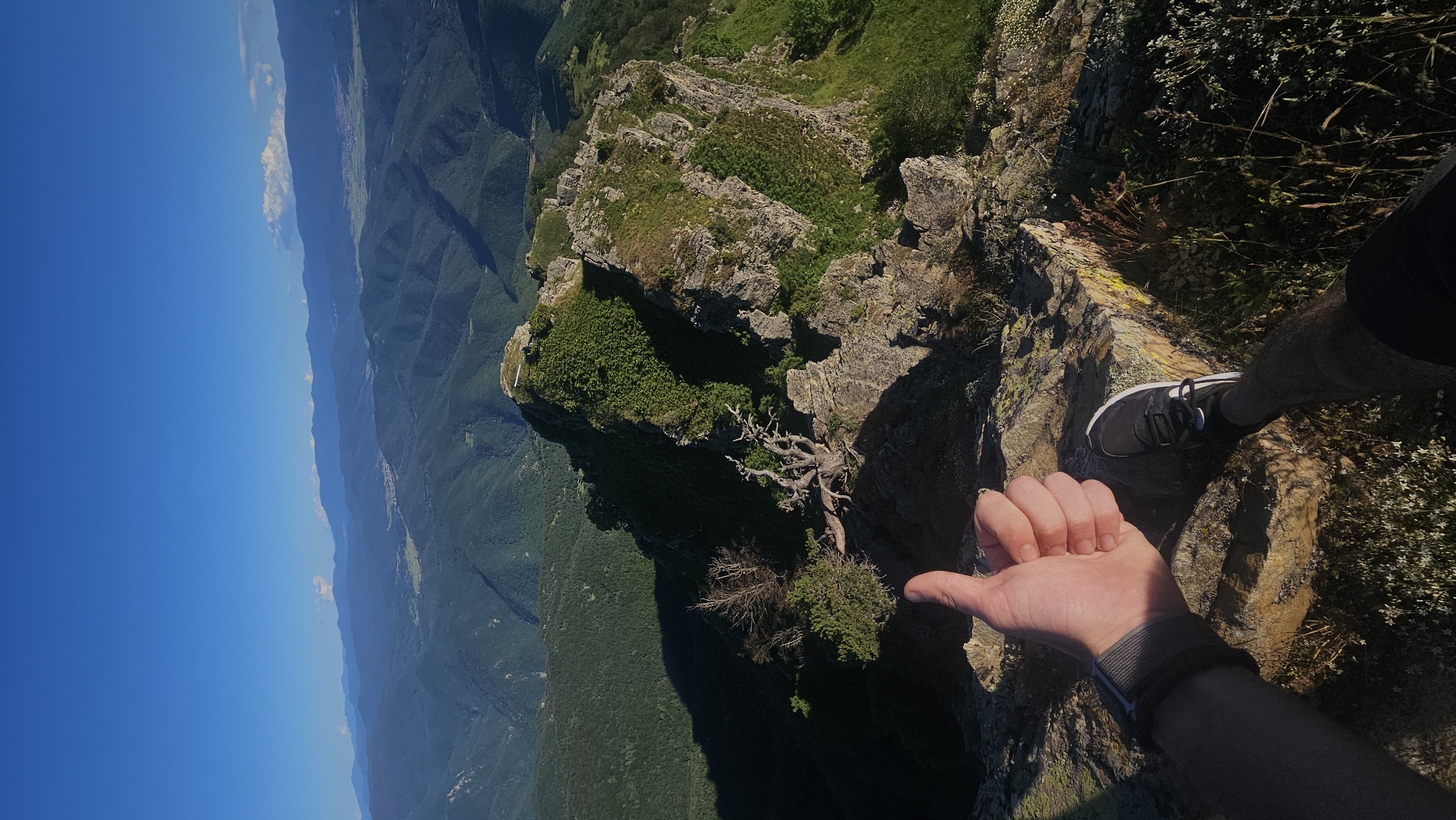The image depicts a person's hand giving a thumbs-up gesture from the edge of a cliff. The perspective is from the person looking down and towards the horizon, with their right hand and a portion of their right leg visible in the foreground. The person is wearing dark shorts and black athletic shoes. The hand is casually extended, thumb up, in a classic gesture of approval or well-being.
The scene is a dramatic mountainous landscape. Steep, rugged cliffs covered in green vegetation drop off sharply into a vast valley. The valley below is densely forested with rolling hills stretching into the distance, creating layers of depth. The sky is a clear, bright blue, suggesting fair weather and a sunny day. In the far distance, a small village or town can be seen nestled within the valley. The time of day appears to be midday, given the bright sunlight and the shadows cast by the cliffs.
The overall mood is one of adventure, freedom, and accomplishment. The precarious position on the cliff edge, combined with the expansive view, evokes a sense of awe and exhilaration. The location appears to be a popular hiking or climbing spot, likely in a natural park or mountainous region. The specific location context provided is Simeonovets, Bulgaria, which aligns with the rugged, mountainous terrain. There is no visible text in the image.