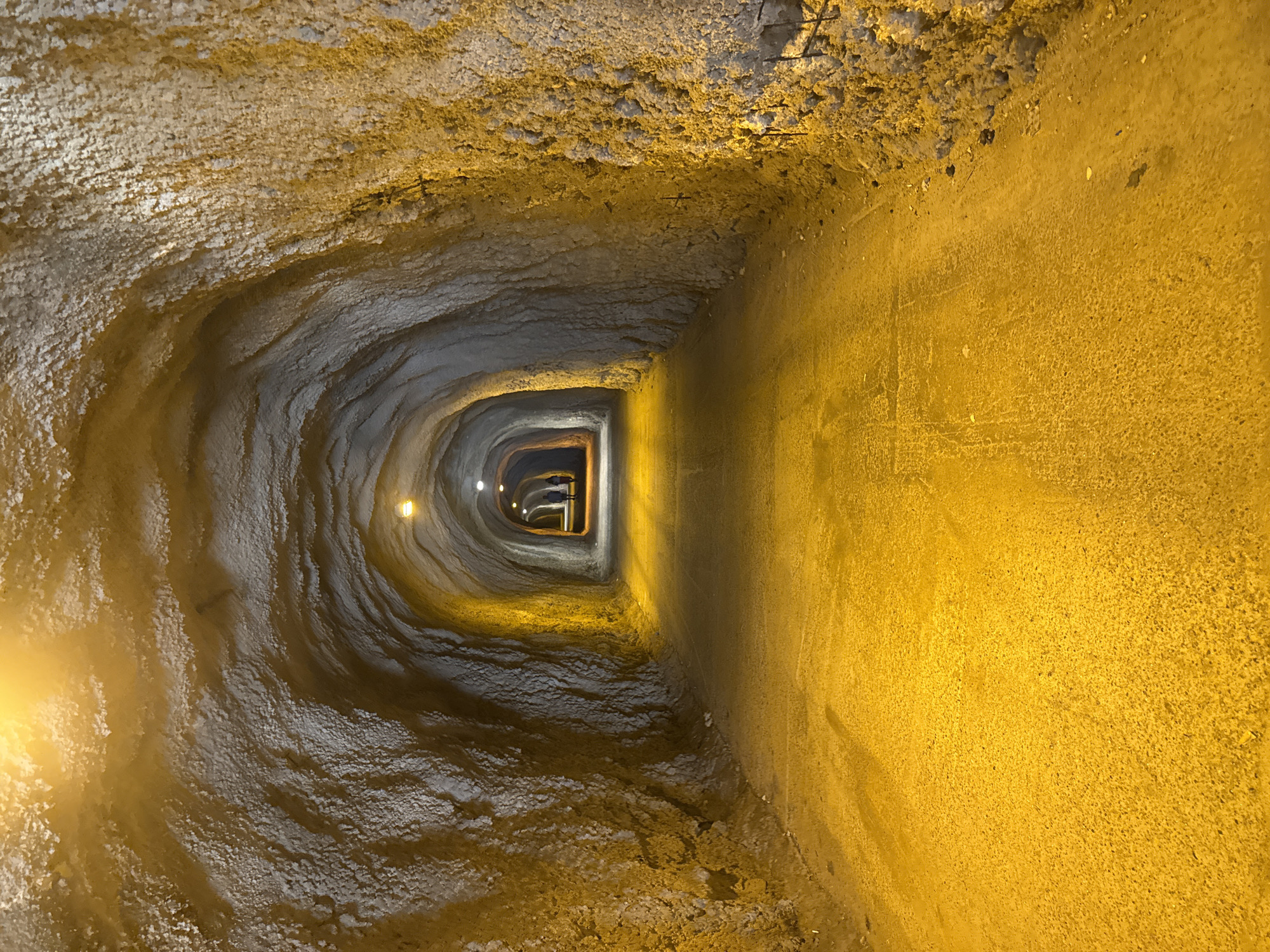 The image shows the interior of a rough, unlined tunnel. The walls and ceiling appear to be made of natural rock or soil, with a textured, uneven surface. Several artificial lights are visible, illuminating the tunnel with a warm, yellowish glow. The floor of the tunnel is a dark, compacted surface, also reflecting the light. In the distance, two indistinct human figures can be seen walking away from the camera. The overall impression is of an underground passage, possibly a pedestrian tunnel or a hiking trail. The provided information states the location is in Kamenovo, Montenegro.