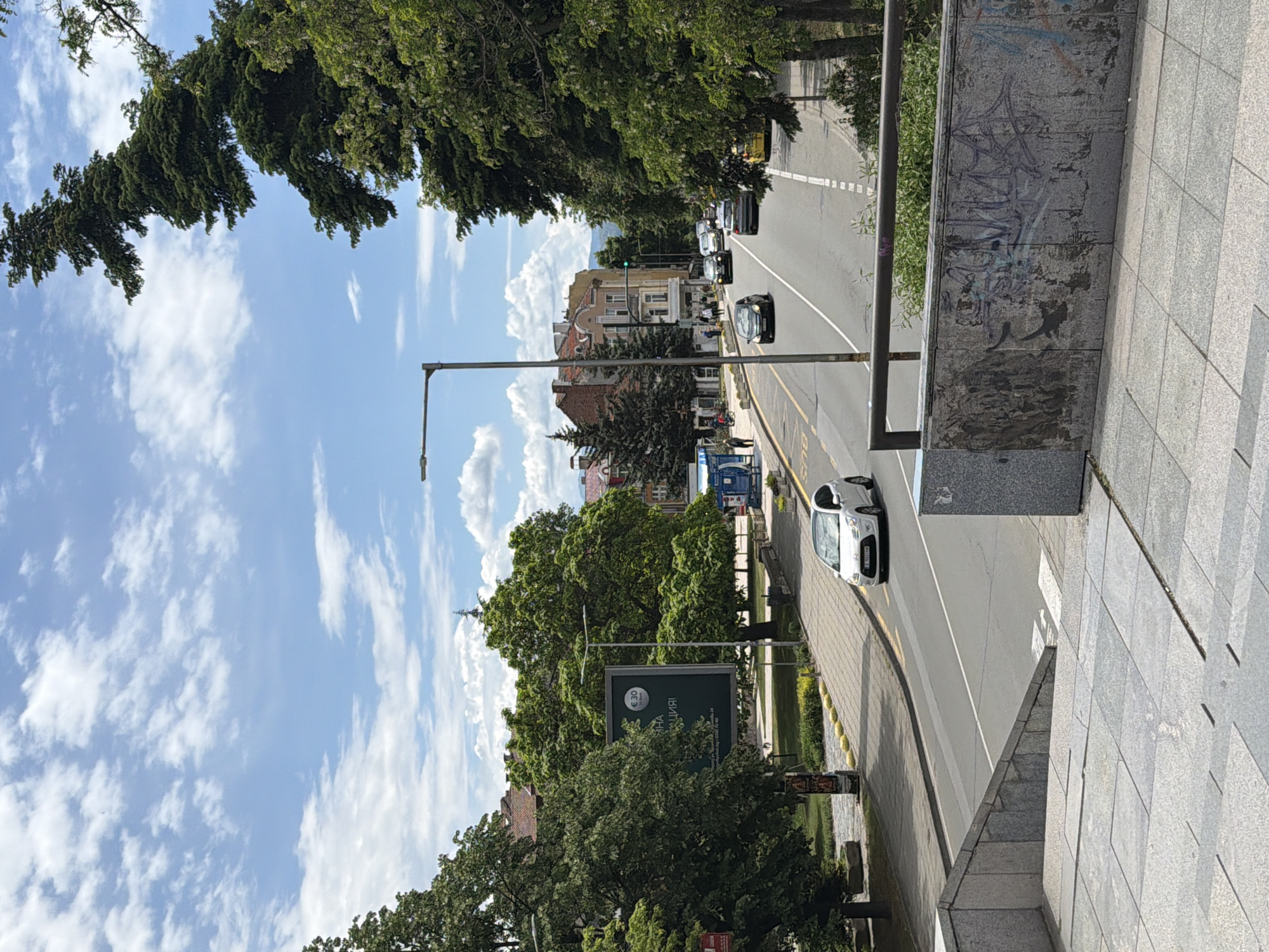 The image shows a street scene in Blagoevgrad, Bulgaria. The camera is positioned on a raised platform or building, looking down on a road lined with trees and buildings. The road is marked with white lines and yellow curbs. Cars are driving on the road in both directions. A lamppost stands on the side of the road. A green billboard is partially visible behind some trees. The sky is blue with scattered white clouds. A wall with graffiti is to the right of the frame.