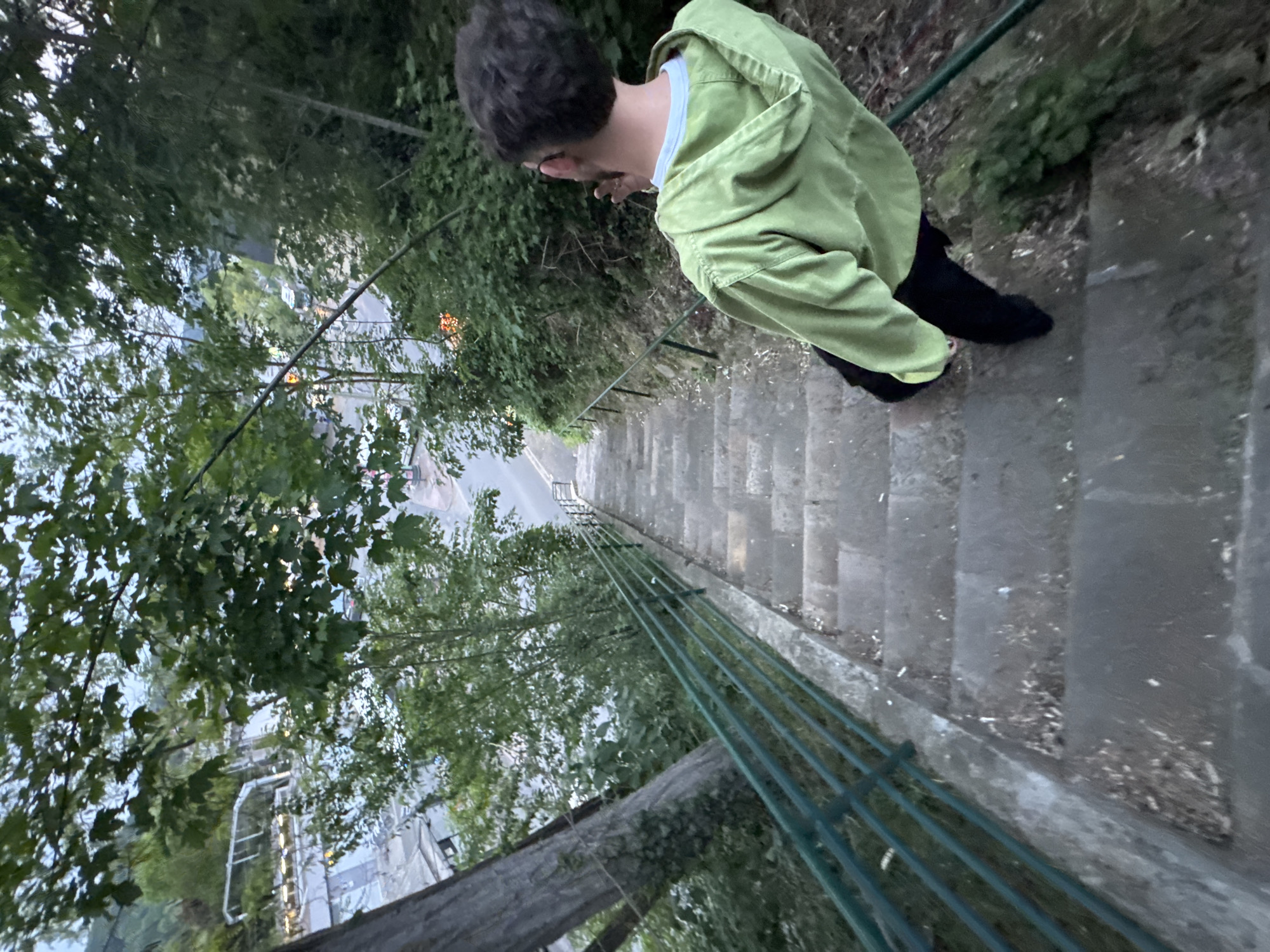 The image depicts a person standing on a long flight of concrete stairs with a green metal railing running alongside. The individual is facing down the stairs, wearing a light green jacket, a white shirt underneath, and black pants. The environment surrounding the stairs is lush with greenery, consisting of trees and shrubbery. In the distance, beneath the trees, hints of buildings and a road can be seen, indicating an urban environment. Based on the location metadata, the image was taken in Orsay, France. The camera angle is canted, giving the impression of a steep descent.