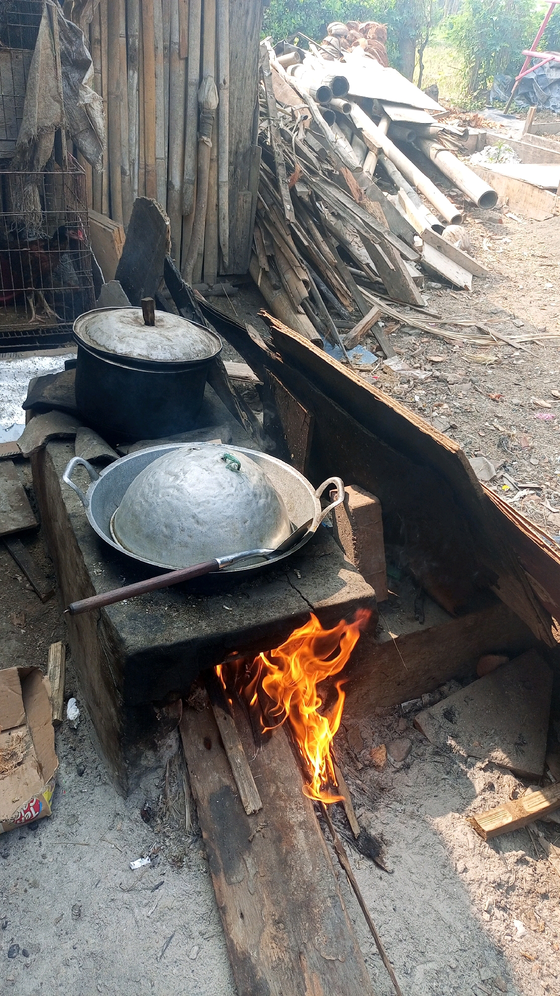In the village of Mangliawan, Indonesia, an outdoor cooking setup is depicted. A makeshift stove, constructed from concrete blocks and wood, is visible. Flames from burning wood are actively igniting beneath a large, inverted metal wok. Inside the wok, a metal pot with a lid is placed, and a serving spoon rests on the edge of the wok. Another, larger pot with a lid is situated behind it, also on a raised platform. To the left, a caged chicken is visible, with parts of what appears to be dried foliage or tarpaulin hanging above it. The background reveals a pile of discarded lumber and cylindrical objects, suggesting a construction or renovation site. The ground is covered with sand and scattered debris. The overall scene conveys a rustic and practical approach to cooking in a rural environment.