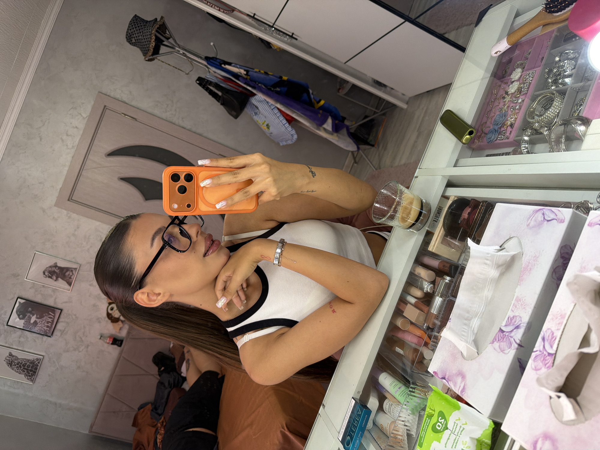 The image shows a woman taking a mirror selfie in a bedroom. She is wearing a white and black tank top and black-rimmed glasses. Her hair is pulled back in a ponytail. She is holding an orange cell phone and has her other hand resting on her chin. On the surface in front of her, there is a makeup organizer filled with various cosmetics, two tissue boxes, a glass with a dark liquid, and a golden electronic device. In the background, there is a bed with a headboard and a partially visible person lying on it. There are also framed pictures on the wall and a door. The scene appears to be indoors, likely a bedroom. The mention of Burgas, Bulgaria, is not supported by visual evidence in the image.