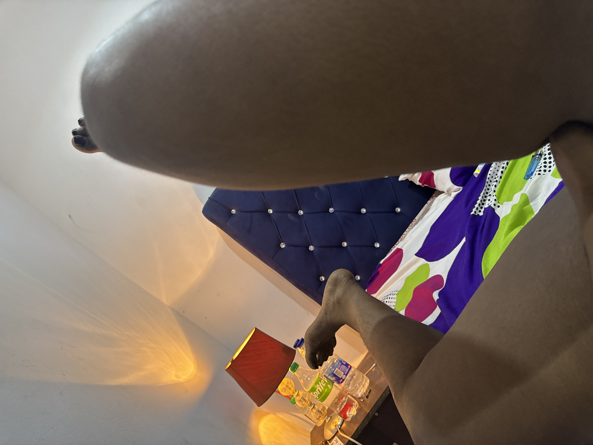 This low-angle photograph captures an indoor bedroom scene, possibly in Abuja, Nigeria, from the perspective of someone lying beneath a person. The foreground is dominated by the dark-skinned legs and feet of an individual, suggesting they are relaxing or lounging on a bed. One foot, visible with dark nail polish on the toes, is positioned towards the upper left of the frame.
In the background, a bed with a prominent dark blue, tufted headboard, embellished with crystal-like buttons, is partially visible. The bed is adorned with a colorful bedspread or duvet featuring a vibrant pattern in shades of purple, green, white, and pink. To the left, a bedside table holds a table lamp with a dark red lampshade, casting warm orange light onto the adjacent light-colored wall. Several plastic bottles are placed on the table, including a green one clearly labeled "Sprite," and other transparent water bottles. The warm, directed lighting suggests the time of day could be evening or a dimly lit interior. The overall mood conveyed is one of relaxation.