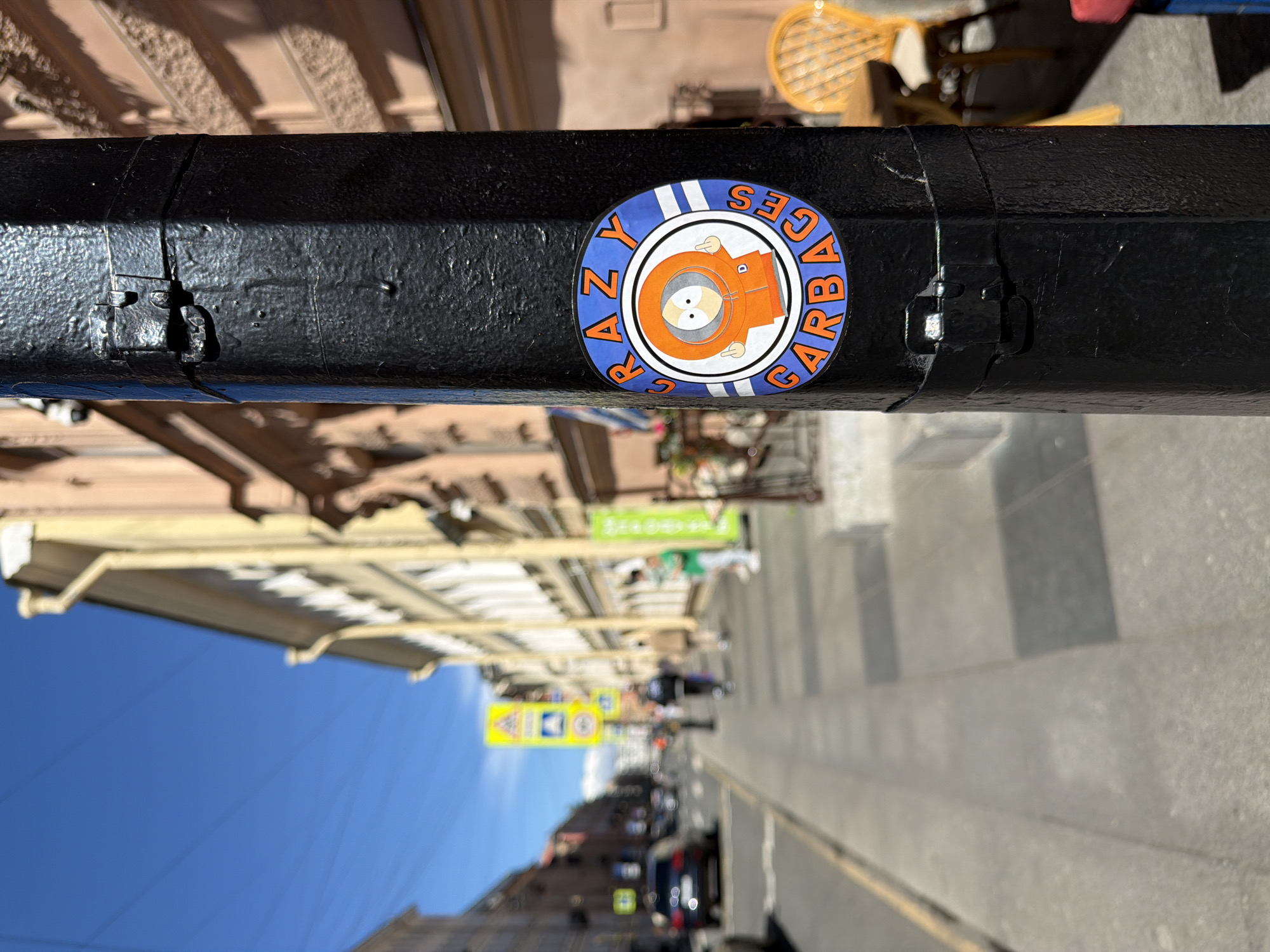 The image depicts a daytime street scene in the city of Saint Petersburg, Russia, under a clear blue sky. In the foreground, prominently centered and in sharp focus, is a black, hexagonal metal pole with a rough texture. A circular sticker is affixed to the pole. The sticker features a blue outer ring with two thin white stripes, containing the orange block letters "CRAZY" at the top and "GARBAGES" at the bottom, each with a white outline. Within this ring is a white circle enclosing a depiction of the cartoon character Kenny McCormick from South Park, wearing his iconic orange parka with the hood pulled up. His hands are visible, each showing a raised middle finger. Below and above the sticker, metal bands with clamping mechanisms are secured around the pole. The pole casts a distinct shadow to its right, indicating bright sunlight from the left.
The background and midground are out of focus, showing a bustling urban environment. To the left of the pole is a paved sidewalk, followed by an asphalt road. Several blurred figures of people are visible walking along the sidewalk in the distance, and blurred vehicles, including a dark-colored sedan, are on the road. Multiple yellow diamond-shaped street signs with black pictograms, characteristic of Russian road signage (e.g., pedestrian crossing signs), are visible further down the street. On the right side of the street, a row of multi-story buildings with light-colored, possibly stucco, facades extends into the distance. Some green vertical banners are attached to the buildings, likely indicating storefronts. A yellow wicker chair and a small table are partially visible on the far right, suggesting an outdoor seating area. Electrical wires are faintly discernible against the bright blue sky above the buildings.
