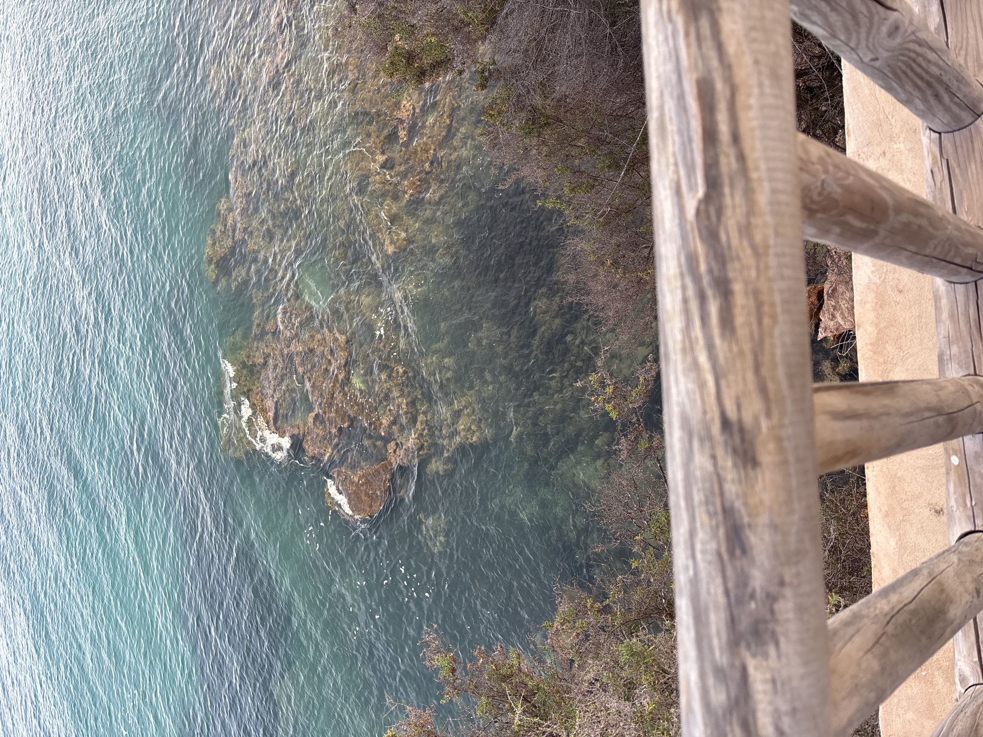 The image shows a view from a wooden railing looking down at the clear turquoise sea near Orihuela, Spain. Rocks are visible beneath the surface of the water, and some patches of vegetation are present on the coastline. The wooden railing in the foreground is weathered and rustic.