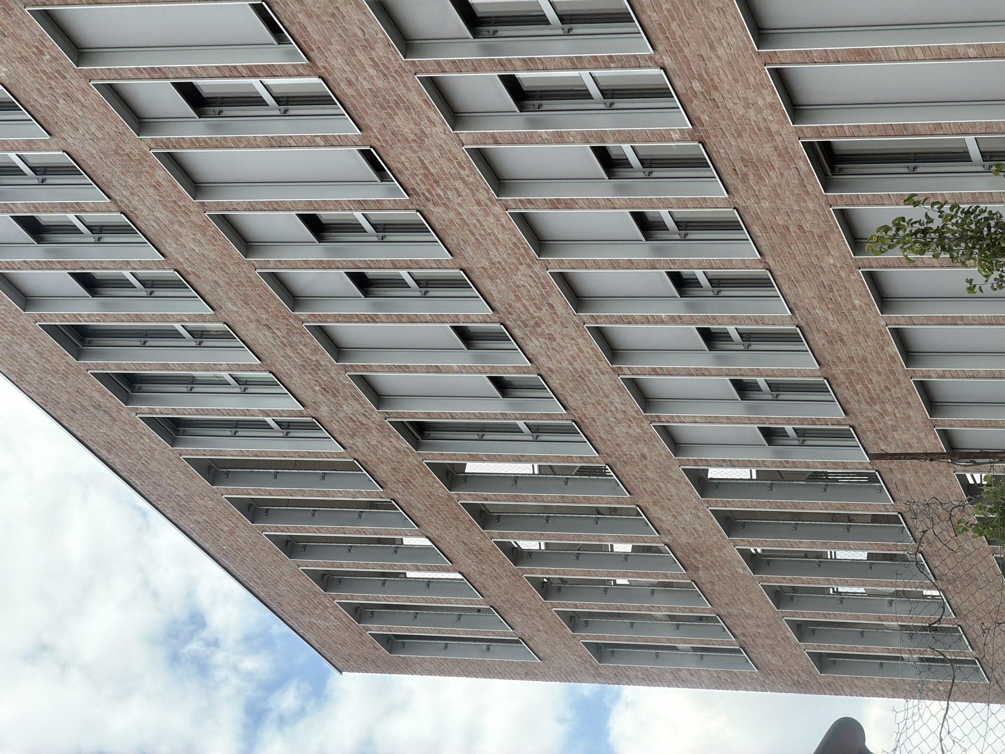 This image, captured in Ivry-sur-Seine, France, offers a unique perspective looking upwards at the underside of a modern building. The building's facade is constructed with reddish-brown bricks, and it features a repeating pattern of rectangular openings, possibly balconies or recessed windows. Each opening is lined with sleek, metallic frames and contains horizontal metal beams, suggesting a robust and contemporary architectural design.
The sky visible in the image is partly cloudy, with patches of blue peeking through the white clouds, indicating daytime, likely during fair weather. A small cluster of green leaves from a nearby tree is visible on the right side, adding a touch of nature to the urban environment. A section of chain-link fence is also present on the right, partially obscuring the view of the building and suggesting a boundary or a protective barrier.
The camera angle is dramatic, emphasizing the geometric repetition and scale of the building's structure. The overall impression is one of modern urban architecture against a natural sky. There is no visible text in the image. The perspective is abstract and focuses on the architectural details rather than people or specific activities.