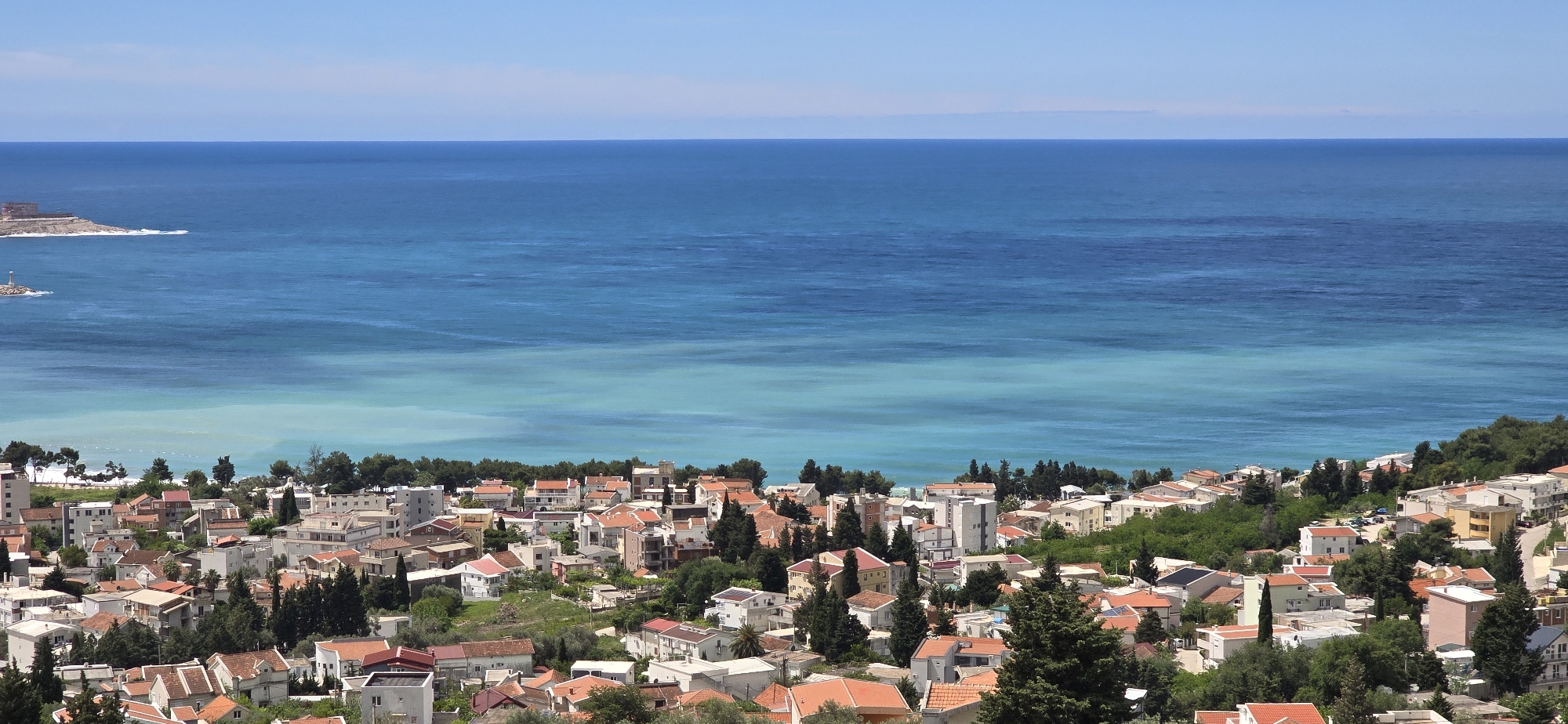 The image shows a high-angle, daytime view of Šušanj, Montenegro. The foreground is dominated by densely packed buildings, most with orange or red tile roofs, interspersed with trees and greenery. Beyond the town, the Adriatic Sea stretches to the horizon, transitioning in color from a turquoise hue near the coast to a deeper blue further out. A small rocky peninsula with a building and what appears to be a lighthouse or similar structure is visible to the left of the image, extending into the sea. The sky is clear blue, indicating good weather. There are no people visible in the image, and no explicit actions are discernible. The environment suggests a coastal town with a mix of residential architecture and natural elements.