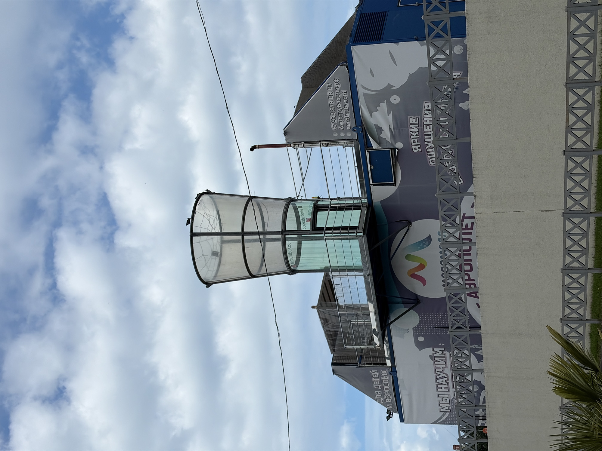 The image shows an outdoor facility in Sochi, Russia, likely an indoor skydiving center. The structure is a multi-story building with a prominent glass cylinder on top, which houses the vertical wind tunnel. The building's exterior is decorated with large graphics and text. One section of text on the side reads "МЫ НАУЧИМ" (We will teach) and "АЭРОПОЛЕТ" (aeroflight), suggesting it is an educational or recreational facility. Another section says "ДЛЯ ДЕТЕЙ И ВЗРОСЛЫХ" (for children and adults). A phone number and website address "+7 938 878 08 07 Aerotrubasochi.rf aerotrubasochi" are also visible on the building, further confirming its identity as an aerotube facility in Sochi. The facility is surrounded by a grey, decorative metal fence and a concrete wall, with some greenery in the foreground. The sky is partly cloudy. There are no people visible in the image.