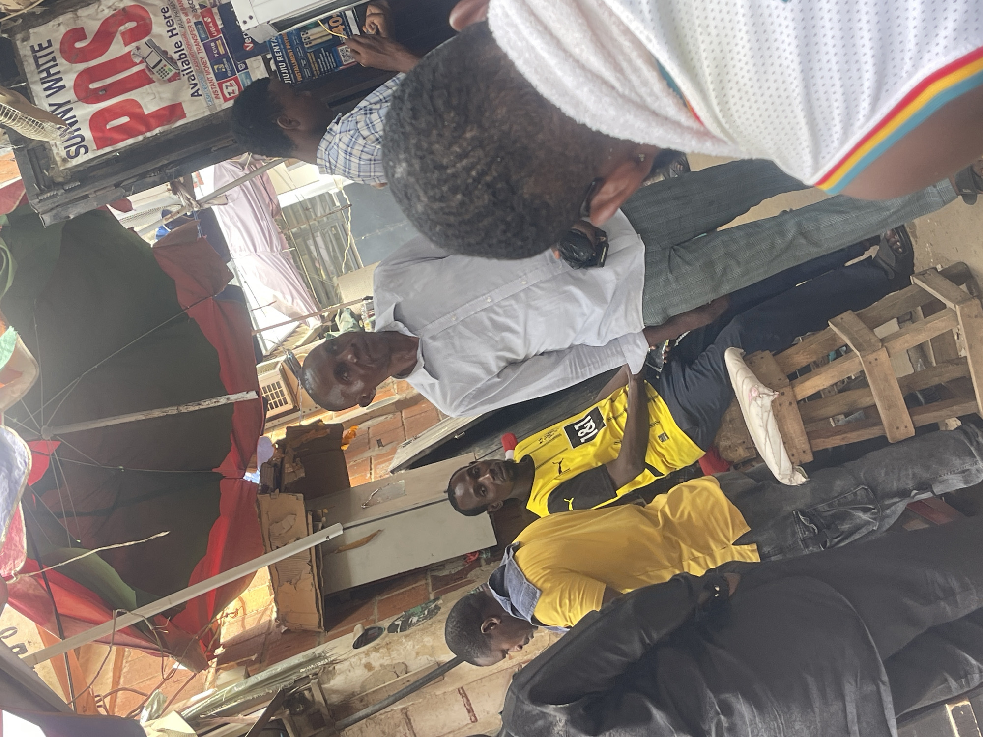 The scene shows an outdoor gathering of people, including four African men, in what appears to be a somewhat cluttered environment. One man is wearing a yellow Borussia Dortmund soccer jersey, another is wearing a white dress shirt, and a third is wearing a yellow shirt with a blue hoodie. Overhead, there are multiple partially opened umbrellas, possibly serving as makeshift shelter. A sign reads "SUNNY WHITE POS Available Here CASH DEPOSIT CASH WITHDRAWAL BILL PAYMENT INSTANT MONEY TRANSFER to all BAN". The location is inferred to be in Abuja, Nigeria.