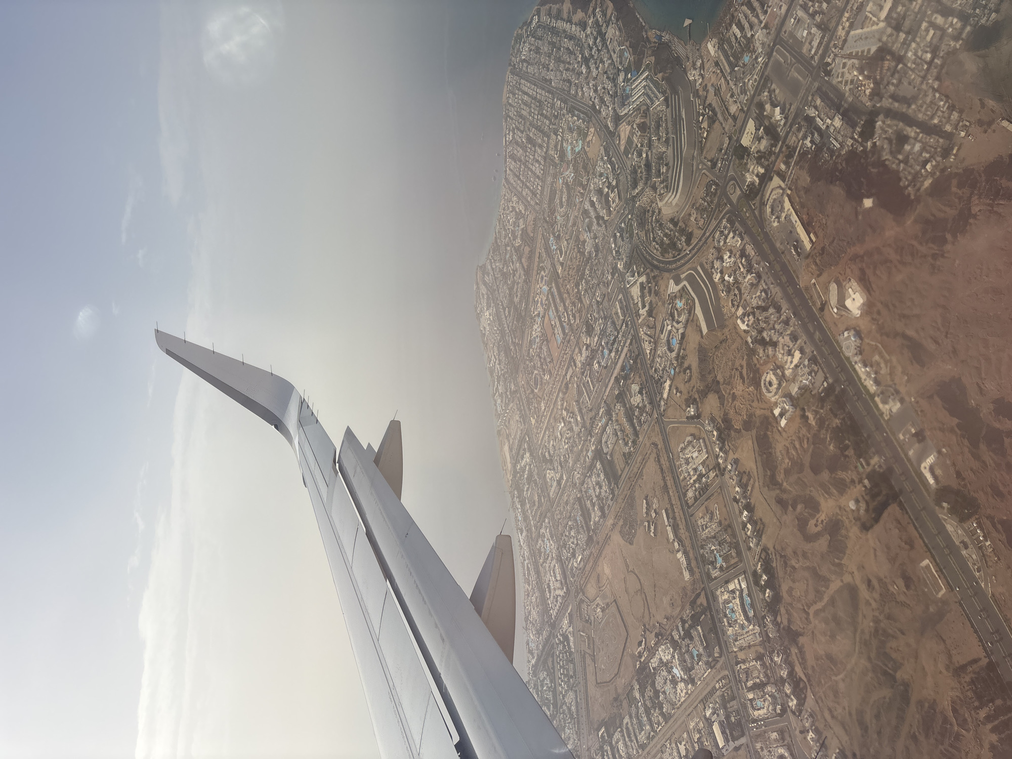 The image shows an aerial view from an airplane window, featuring the plane's wing on the left side of the frame. The environment below appears to be a city or urban area near a coastline. The urban layout is characterized by a dense network of buildings and roads. The area further inland transitions into a more arid, desert-like landscape. The sky is overcast. The location is Unknown, Unknown.
