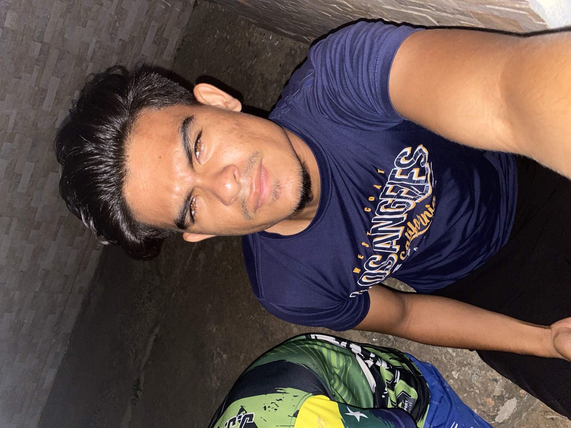 The image is a tilted selfie of a young man, likely taken in a casual, low-light setting in Juan Griego, Venezuela. The man, with dark hair styled back and a neatly trimmed goatee, looks directly at the camera with a neutral to slightly contemplative expression. He has brown eyes and a clear complexion. He is wearing a dark blue short-sleeved t-shirt with "LOS ANGELES CALIFORNIA" and other faint white and orange text, possibly "WEST COAST," printed on the front. He is also wearing dark pants.
The background consists of a dark, textured concrete floor and a partially visible wall made of dark-colored blocks or bricks. The dim lighting suggests the photo was taken at night or in an area with poor illumination. Part of a second individual is visible in the bottom right corner of the frame, wearing a vibrant shirt with green, black, and white patterns, and some indistinct text that appears to be "SPORT." The scene gives the impression of a relaxed, informal gathering outdoors or in a semi-enclosed space.